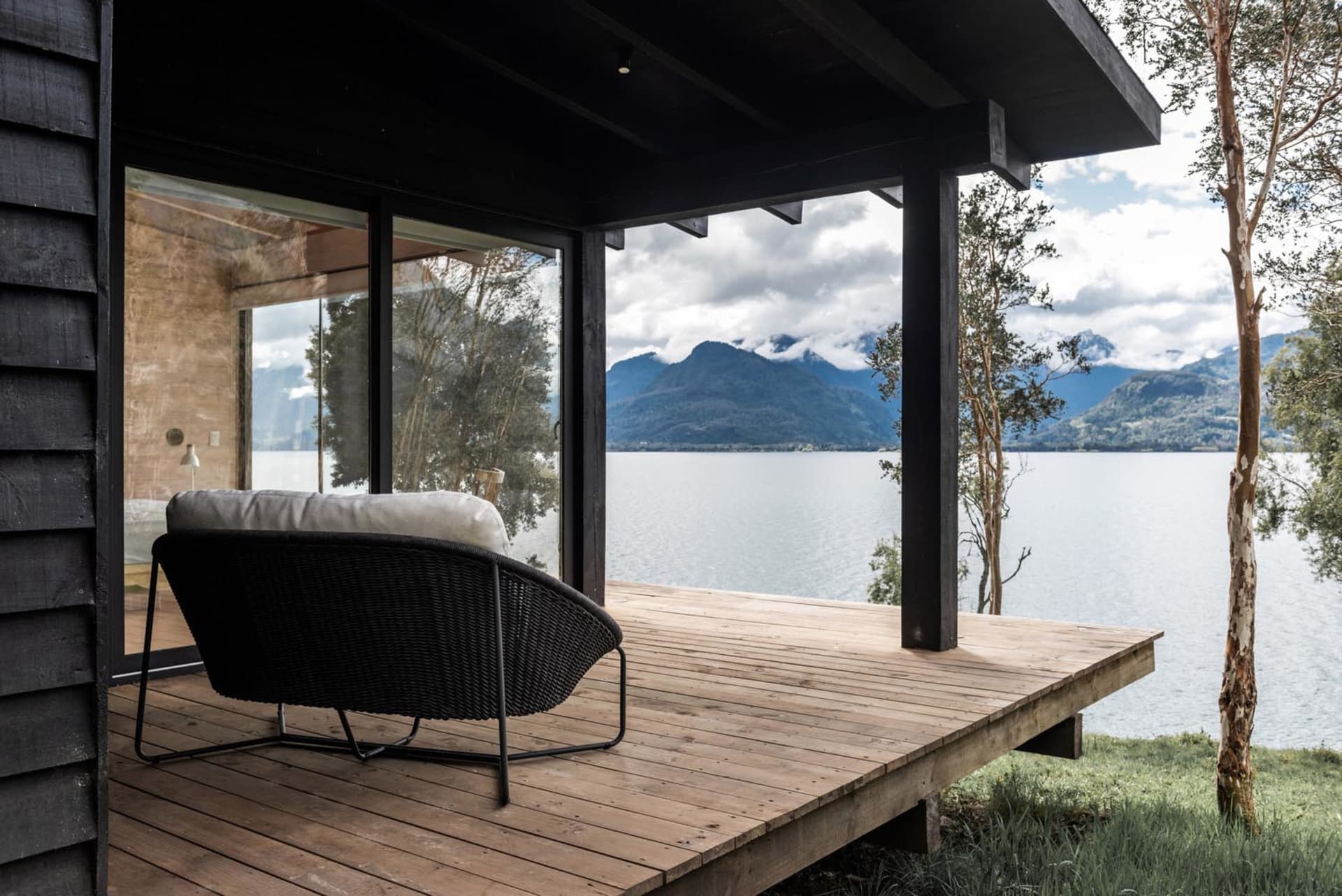 Modern residential deck featuring a wooden platform with a black wicker lounge chair overlooking a serene lake and mountains.
