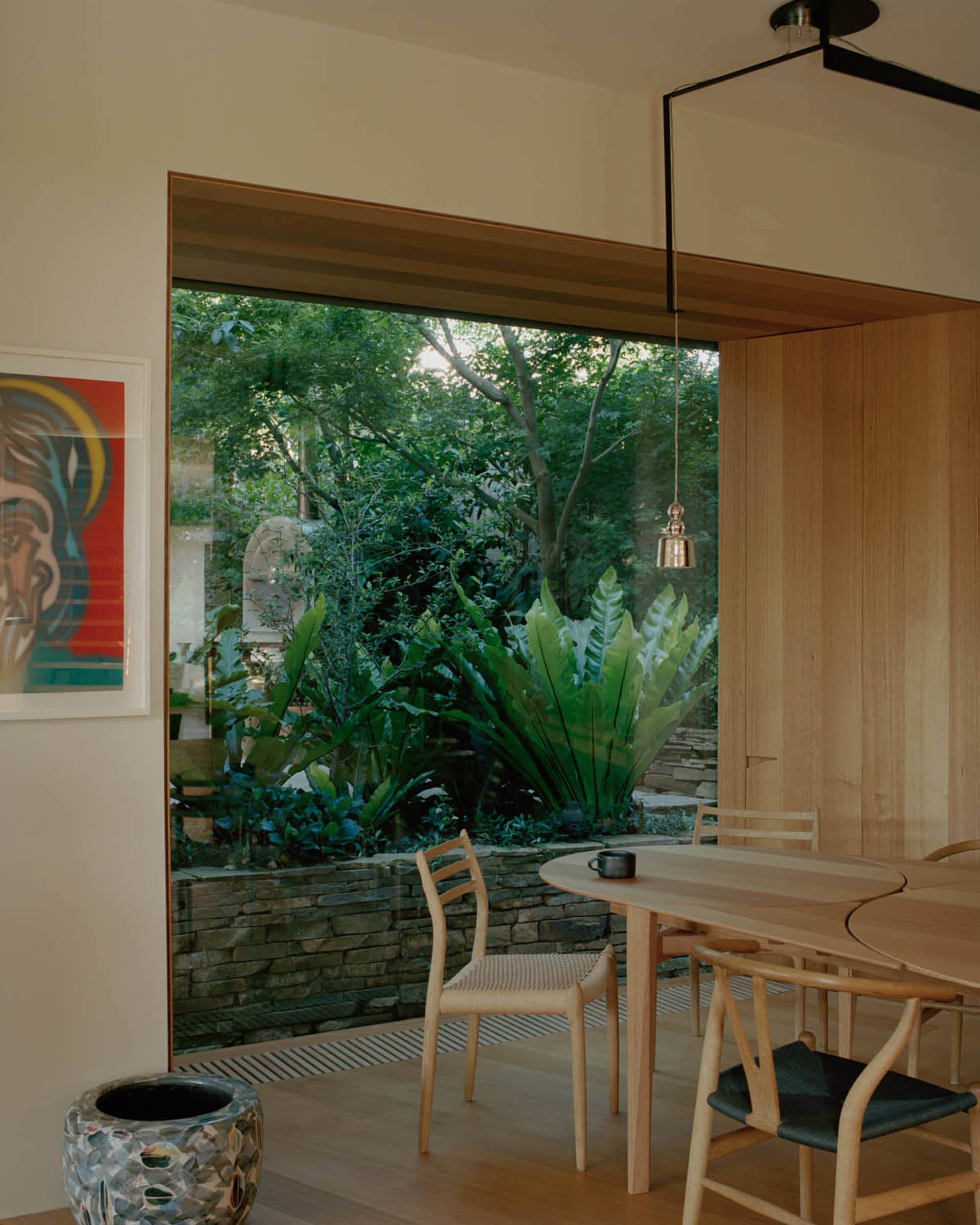 A modern wooden dining room with a large window, greenery outside, and minimalist furniture.