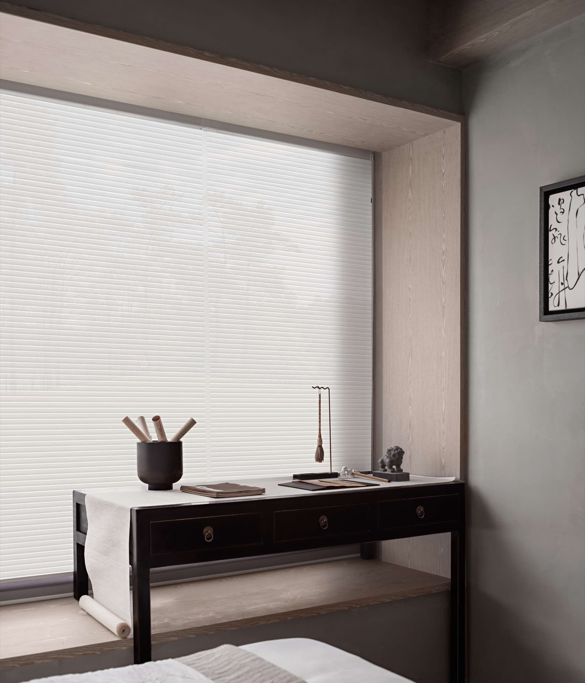 A minimalist bedroom scene with white blinds covering a window, a dark wooden table, and a framed abstract painting on the wa