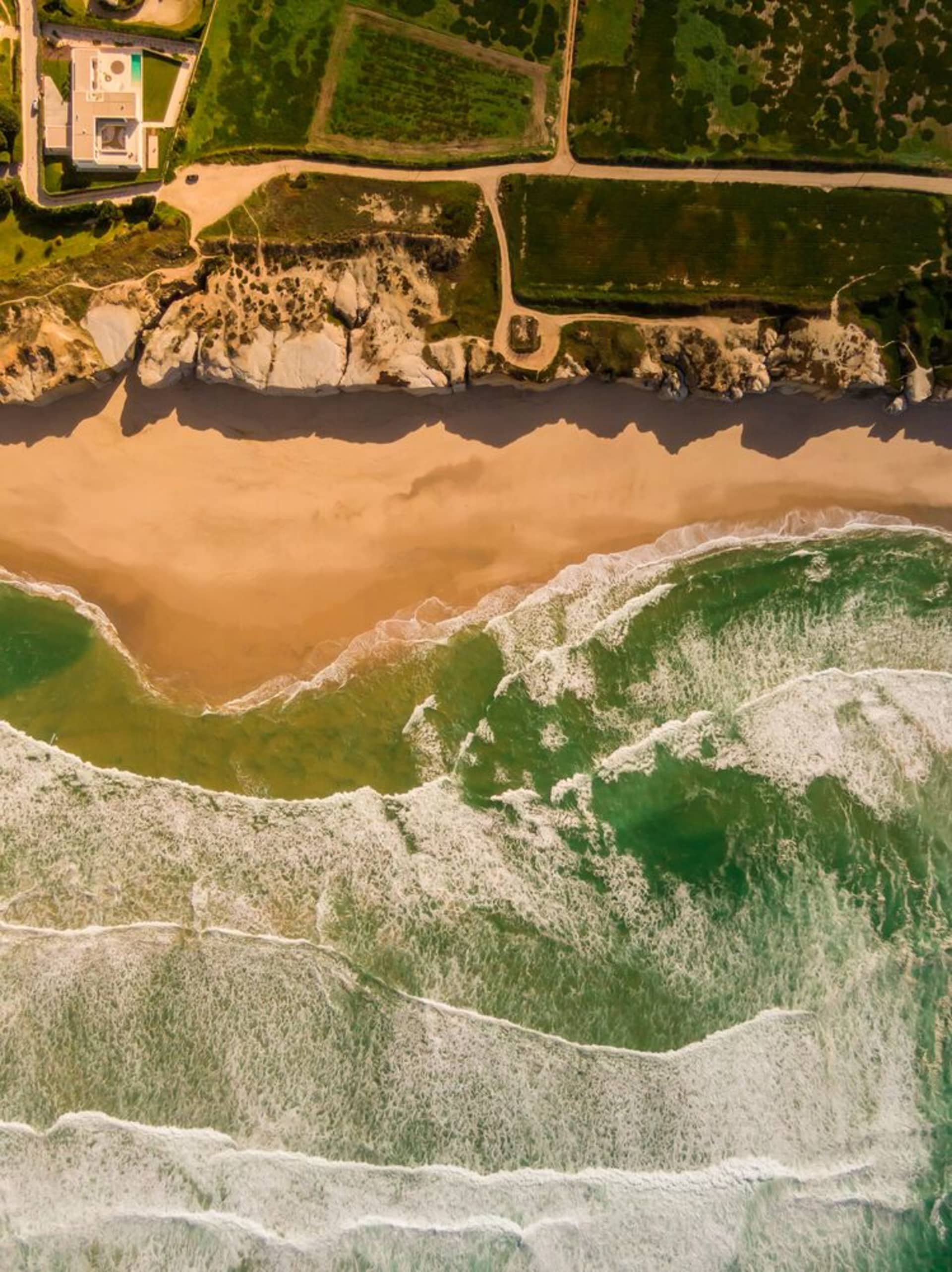 An aerial view of a beach house with a concrete exterior, cobblestone pathways, and a green sea with waves gently crashing ag