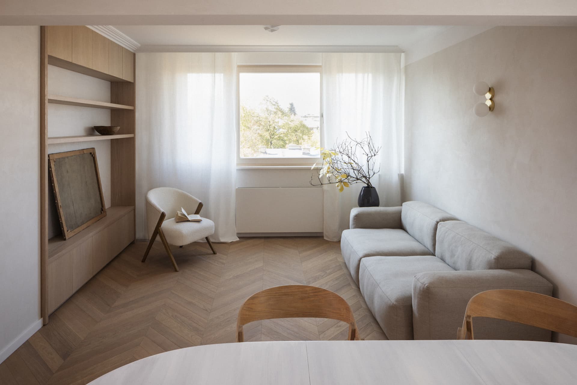 A minimalist living room with a wooden floor and furniture, featuring a white chair, a light-colored sofa, and a bookshelf.