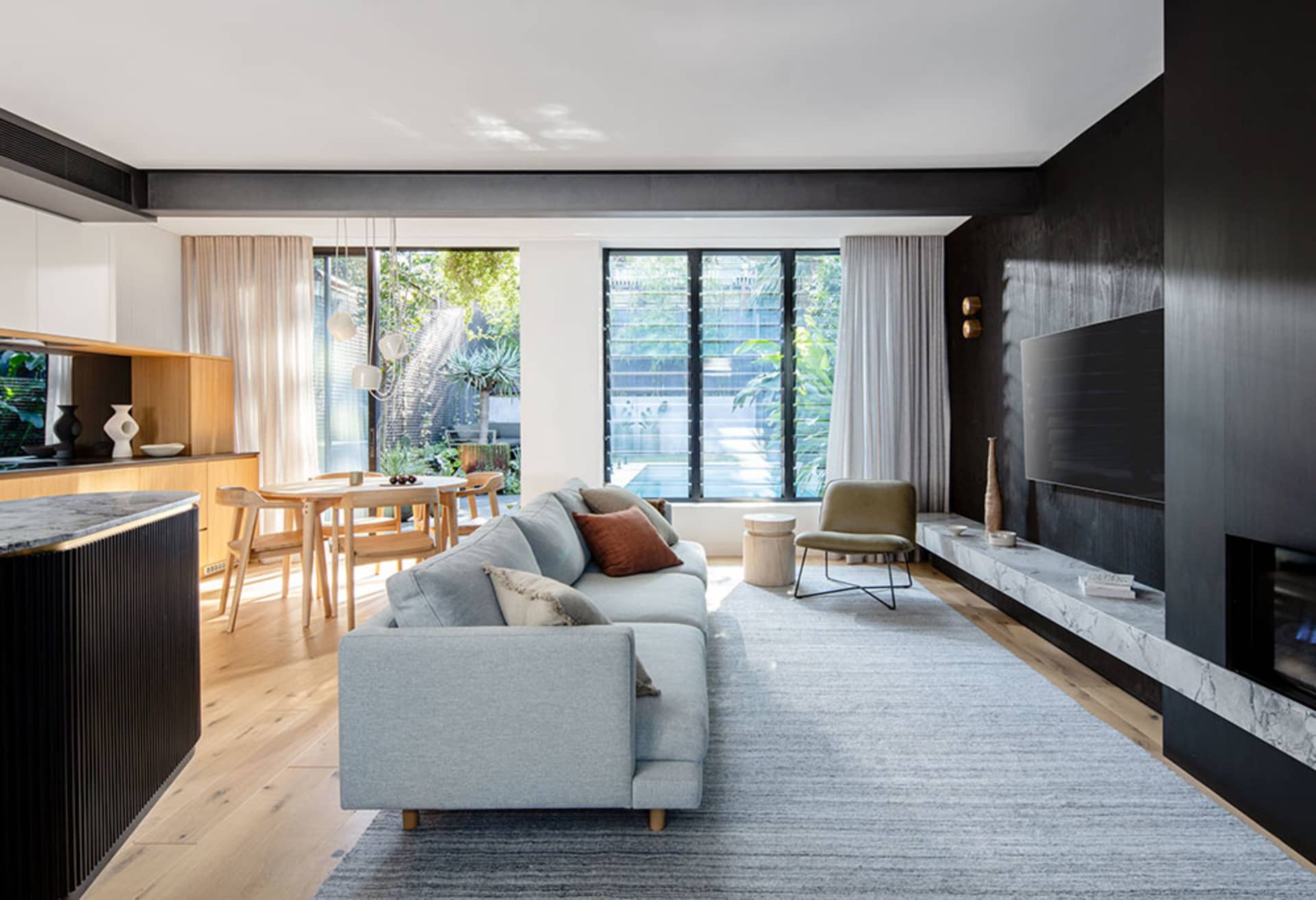 A modern living room with a grey sofa on a grey rug, wooden flooring, a kitchen counter with vases, and large windows overloo