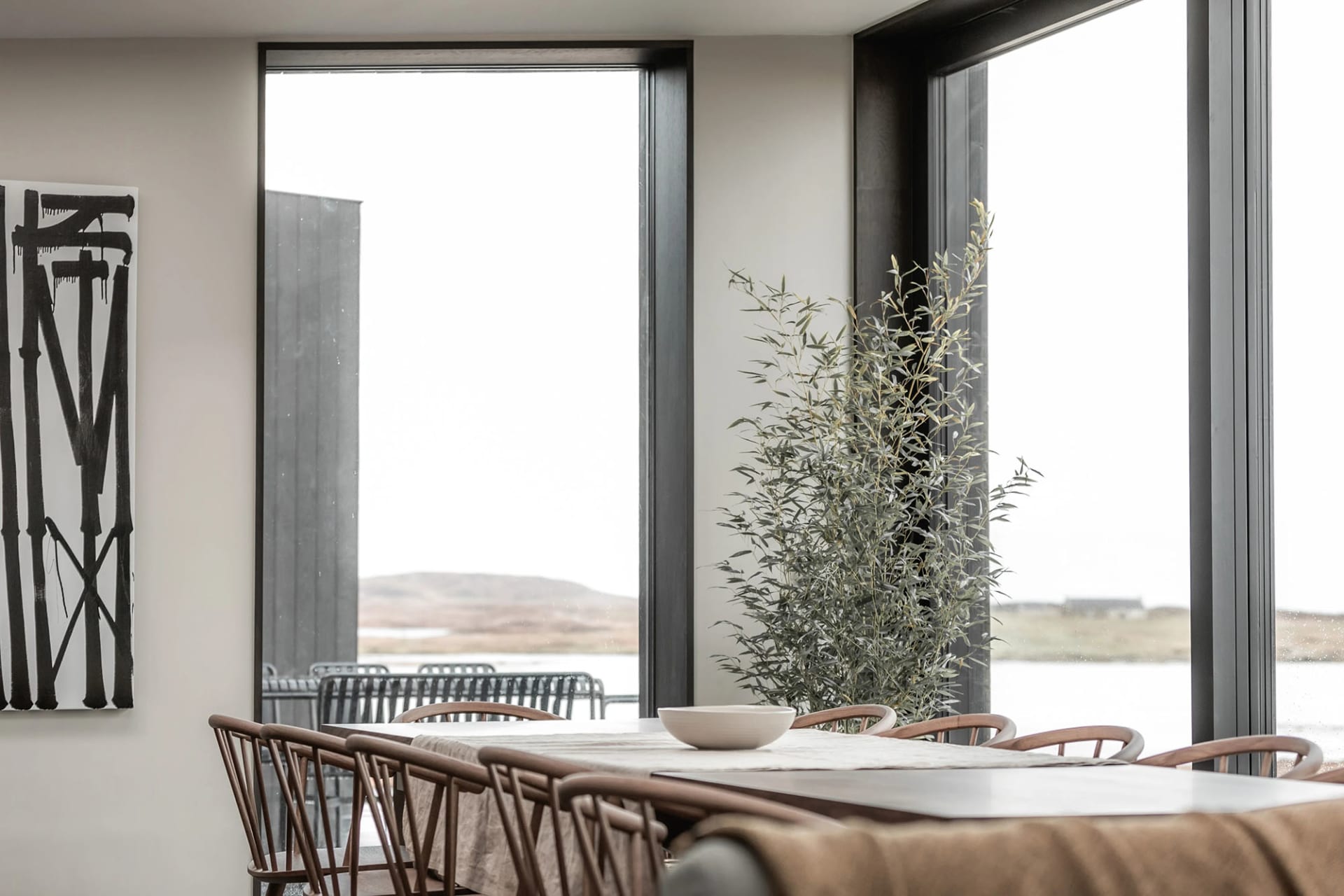 A minimalist dining room with wood furnishings and a large window with greenery outside.