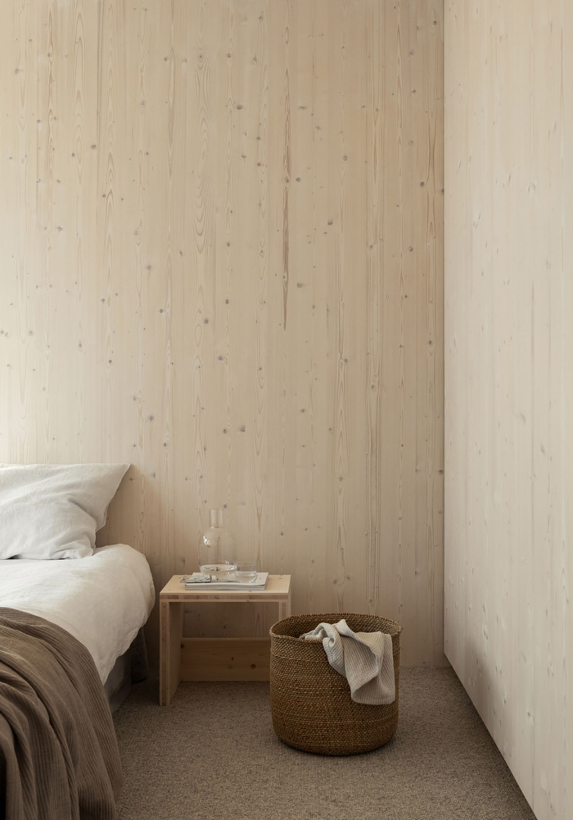 A minimalist bedroom featuring wooden walls and a wooden nightstand with a glass decanter and two glasses.
