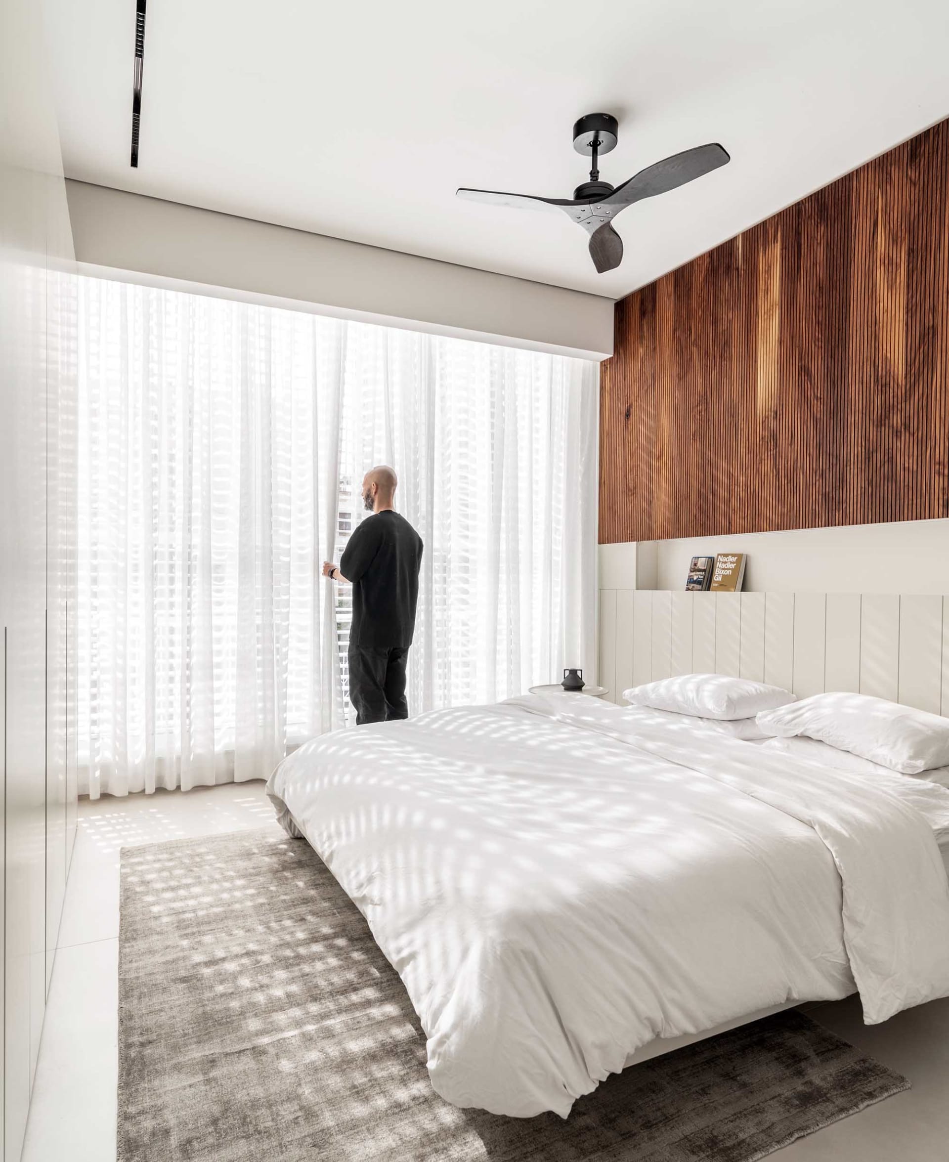 A minimalist bedroom featuring white bedding, a wooden headboard, and sheer curtains allowing sunlight to filter through.