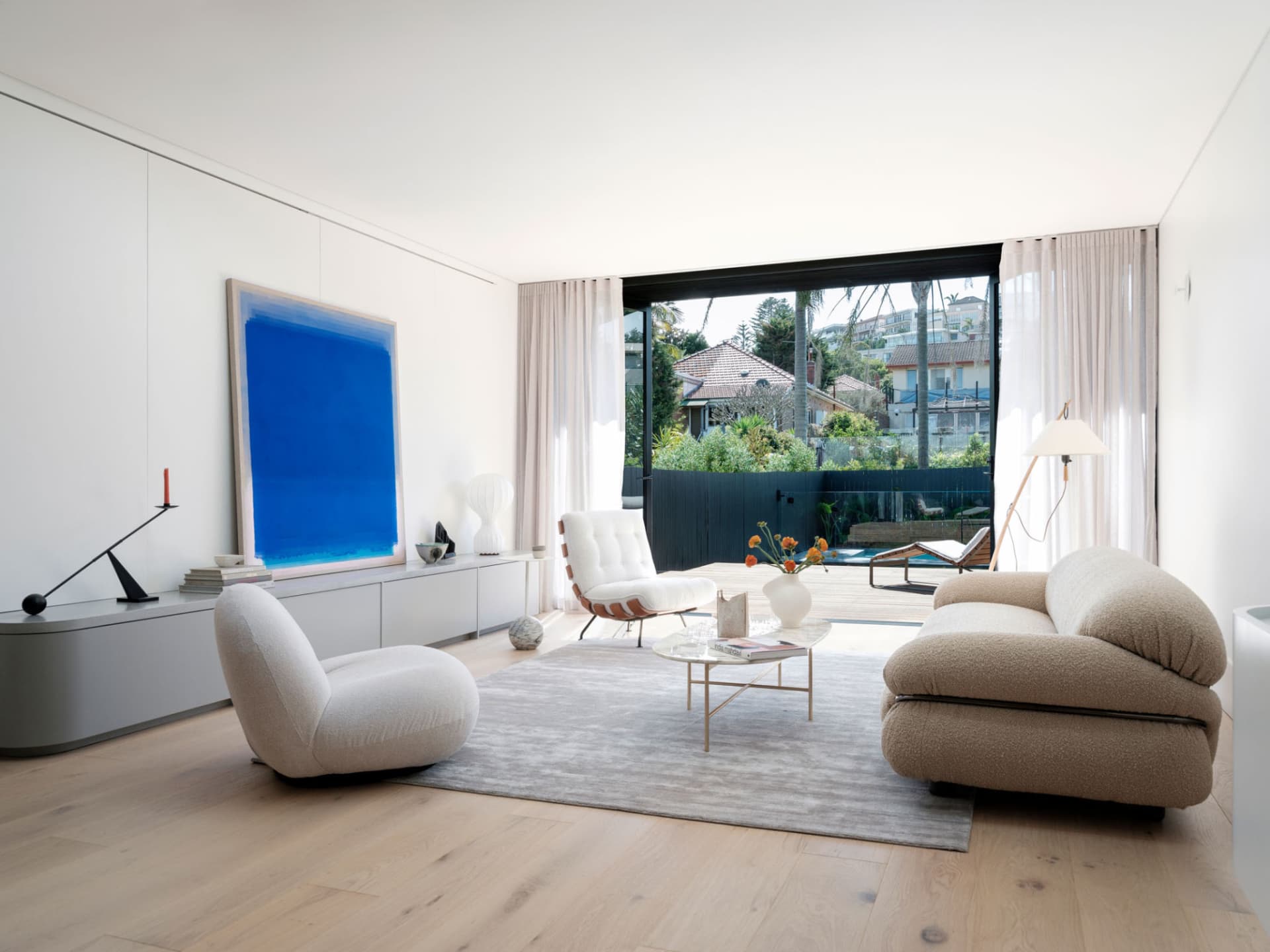 A minimalist living room with modern furniture, a large blue artwork, and a view of the outdoors through large windows.