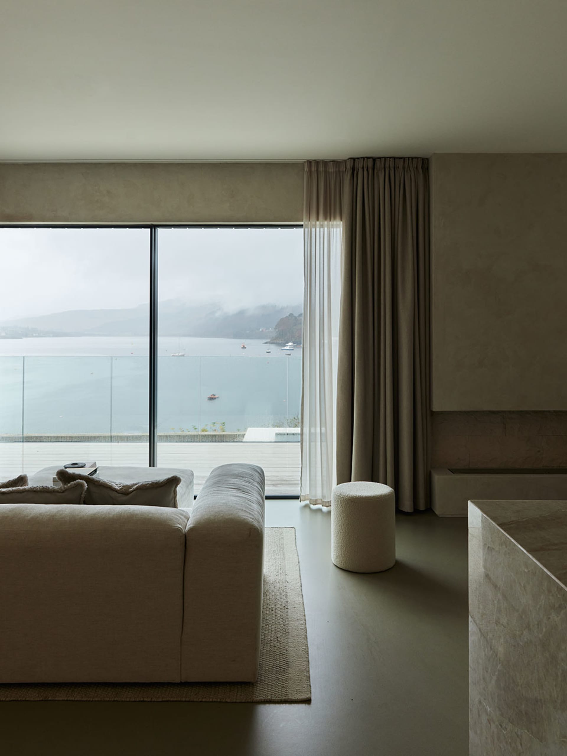 Modern minimalist living room with a large window overlooking the water, featuring a beige fabric sofa and a glass coffee tab