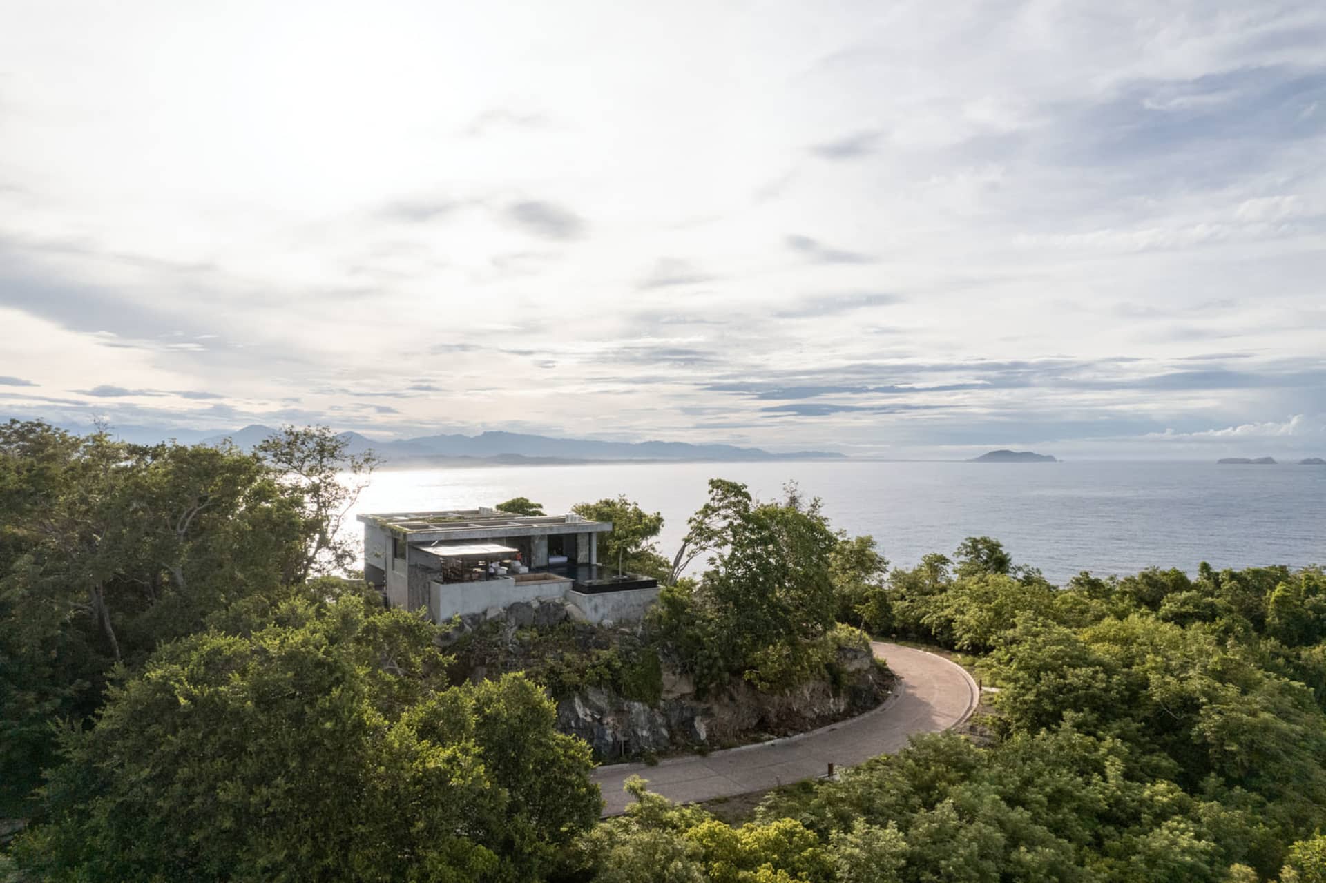 Modern architectural home with concrete walls and a glass terrace overlooking the ocean from a rocky cliffside.