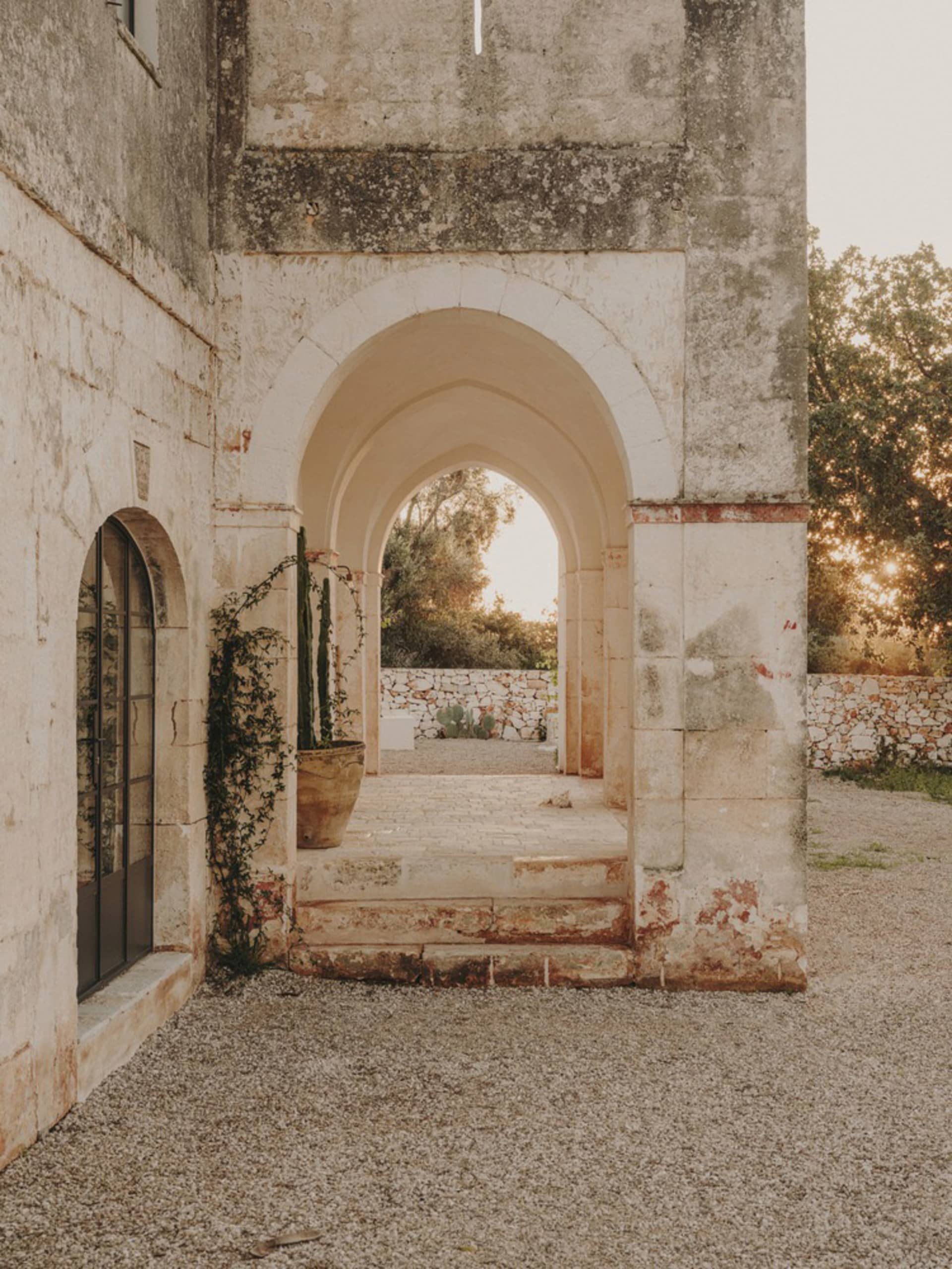 A stone entryway with an arched opening, leading to a residential courtyard with plants and trees in the background.