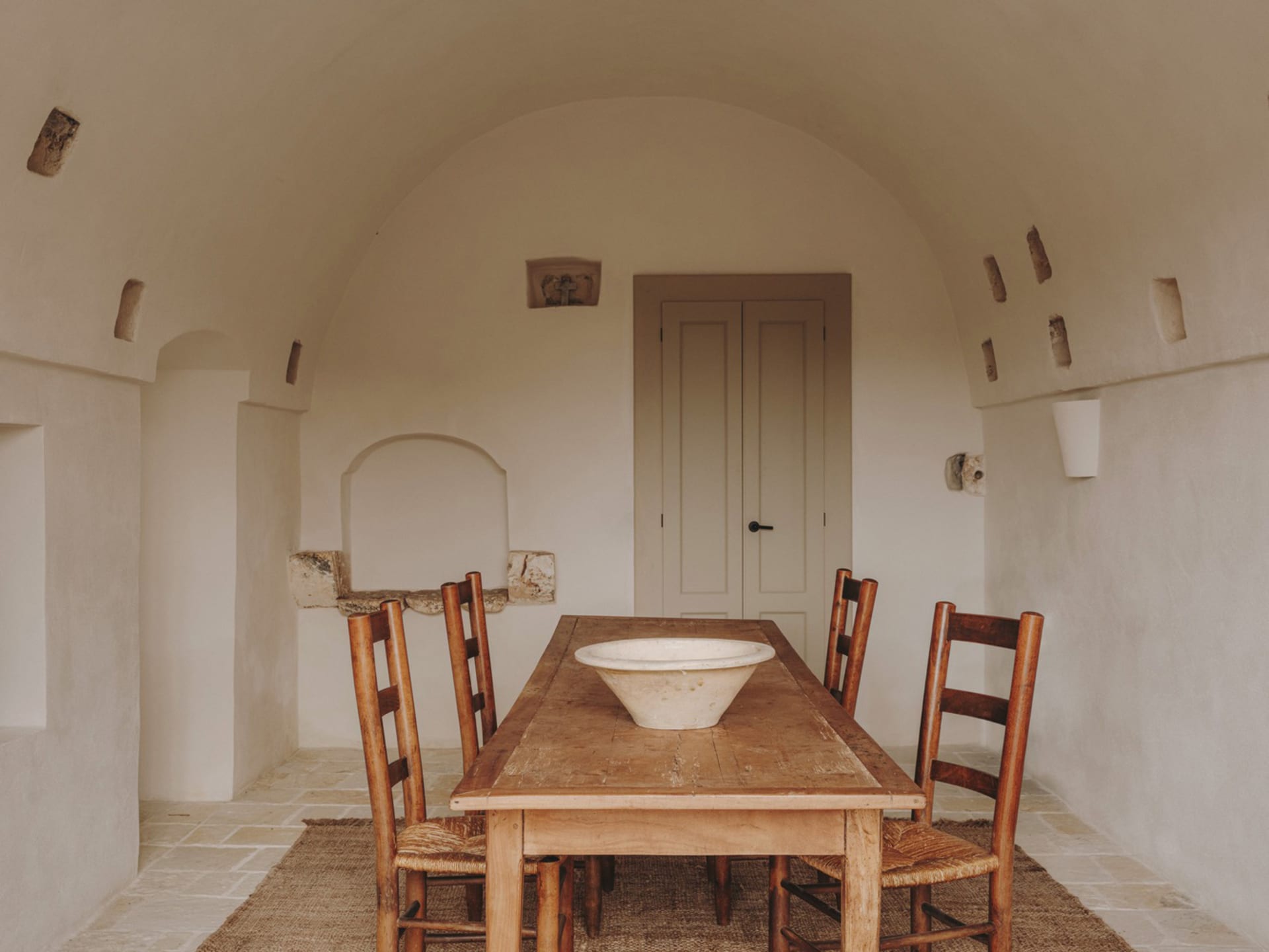 A rustic dining room with a wooden table, chairs, and a central bowl on the table. The walls are made of drywall with visible