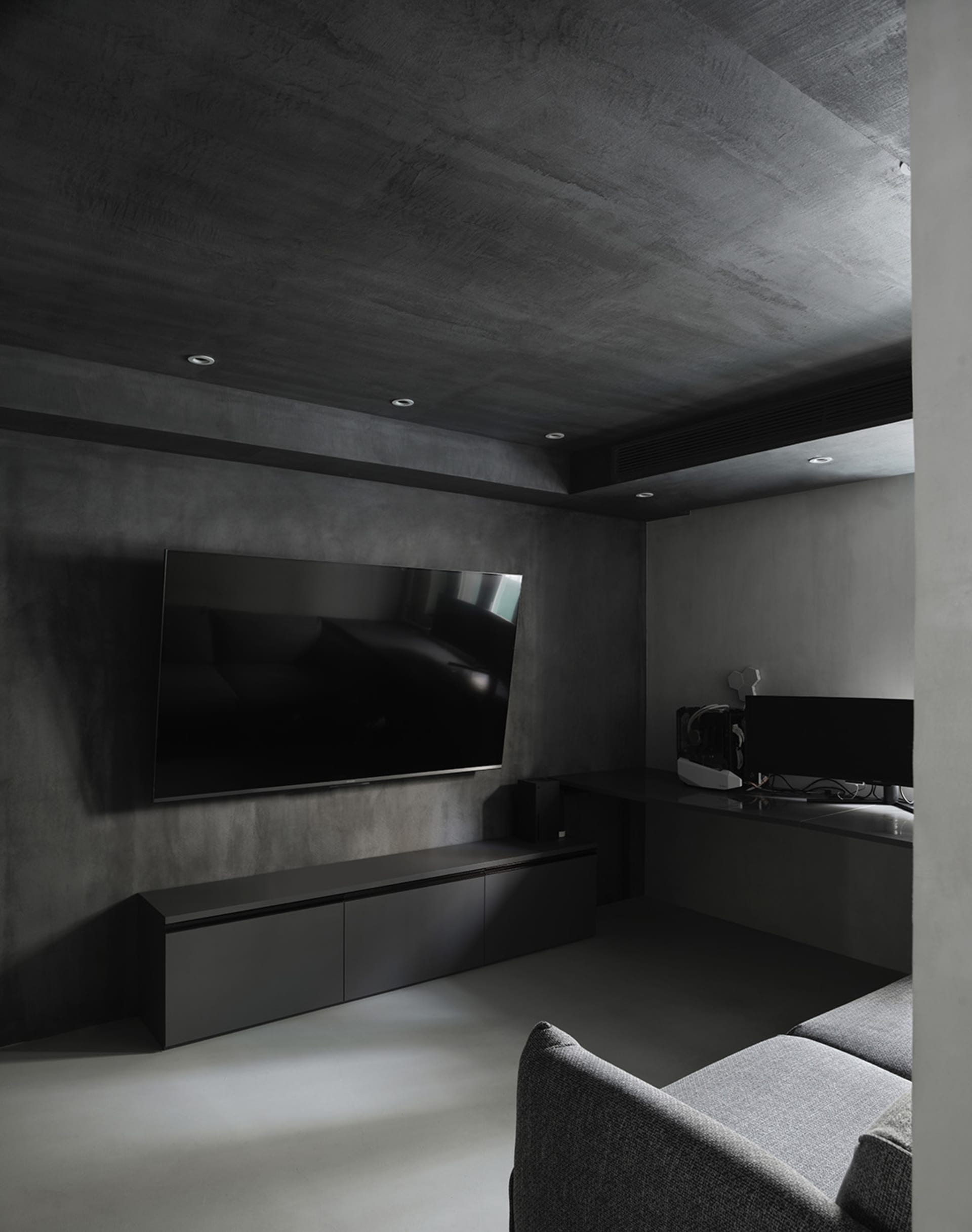 Modern industrial-style living room with concrete walls and floor, featuring a flat-screen TV and grey upholstered furniture.