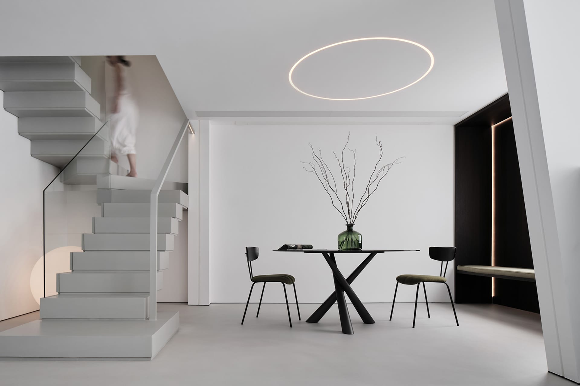 A minimalist dining area featuring a metallic table and chairs, with a circular light fixture and a person descending a stair