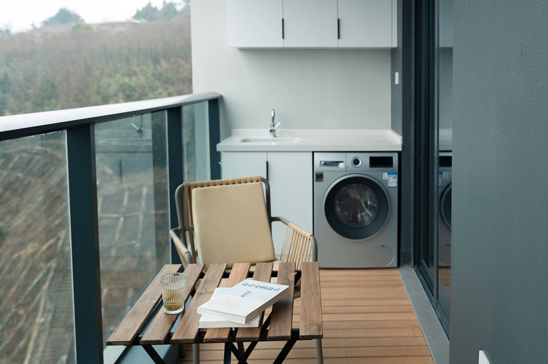 Balcony laundry room with modern wood and metal furniture, glass railing, and white appliances.