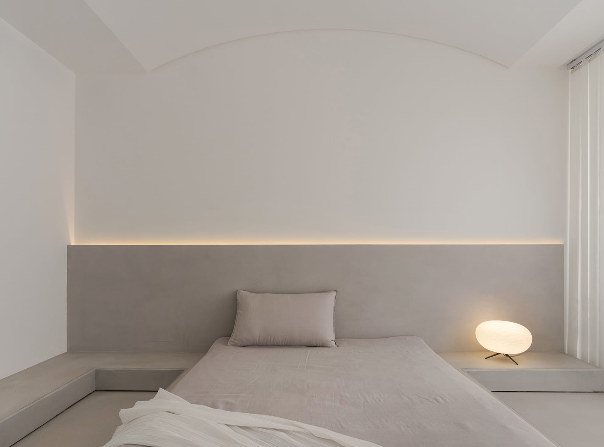 Minimalist bedroom with concrete walls and floor, featuring a bed with simple bedding and a small round lamp.