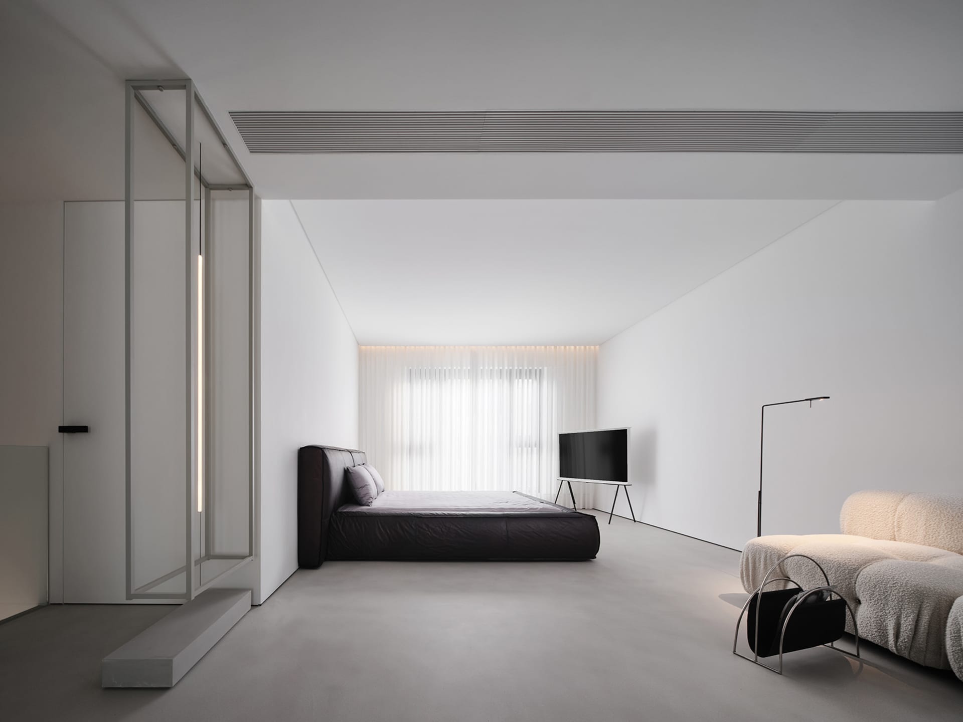 A minimalist bedroom featuring a black bed, a white couch, and concrete flooring, with a floor lamp and a television in the b