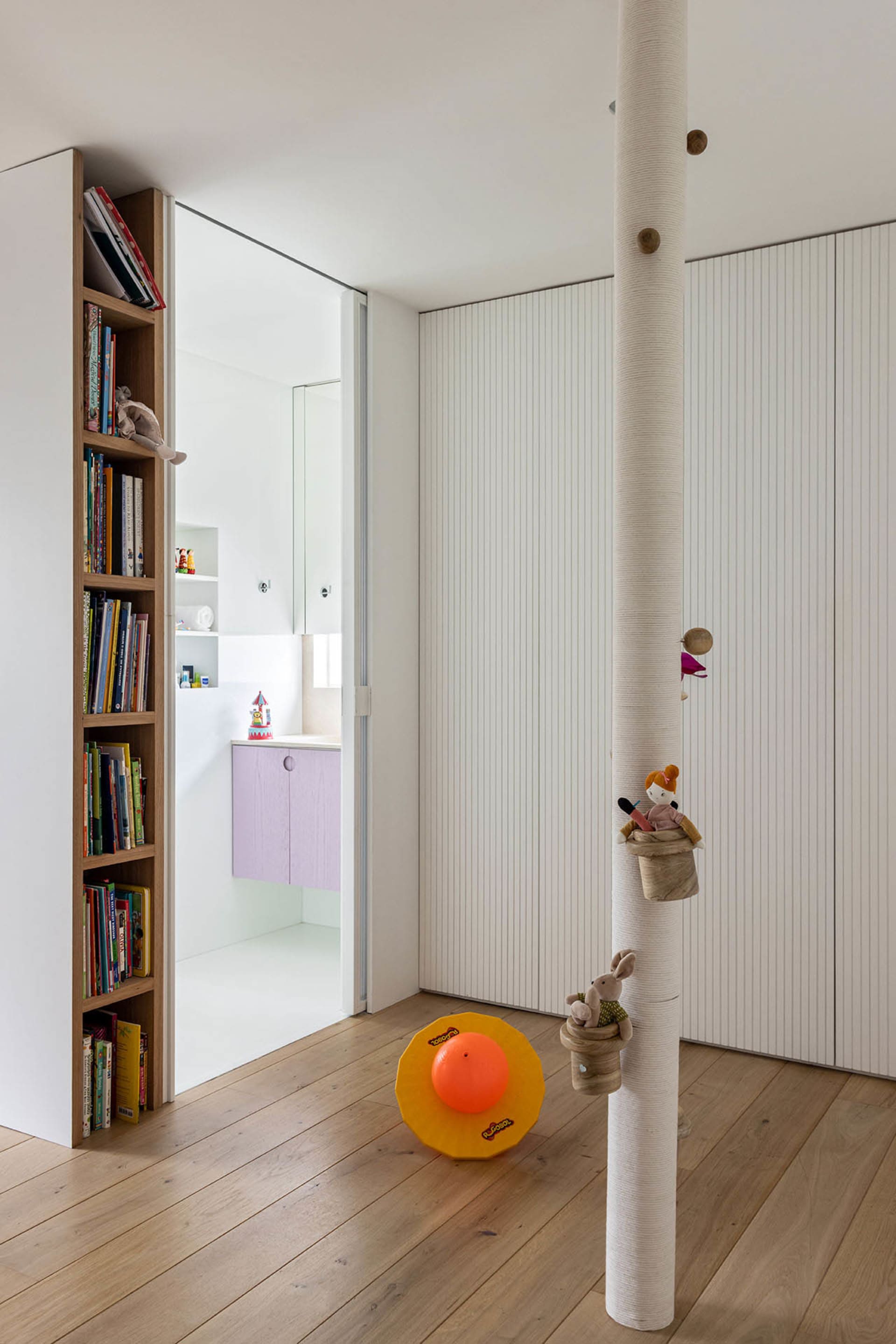 A modern children's room with white paneled walls, wooden shelves containing books and toys, and a wooden floor.