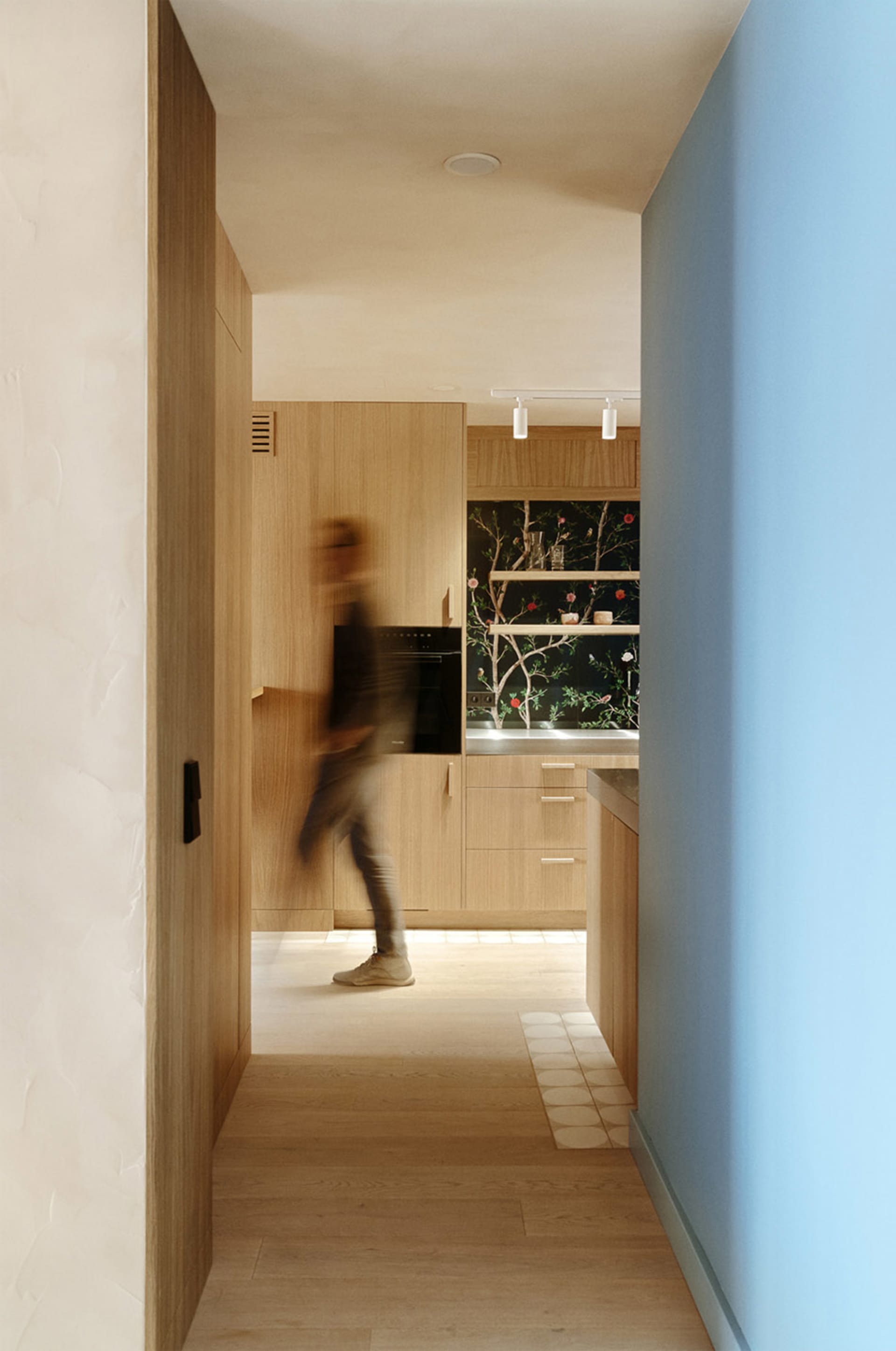 A person walking in a modern kitchen with wooden cabinets and a blue wall.