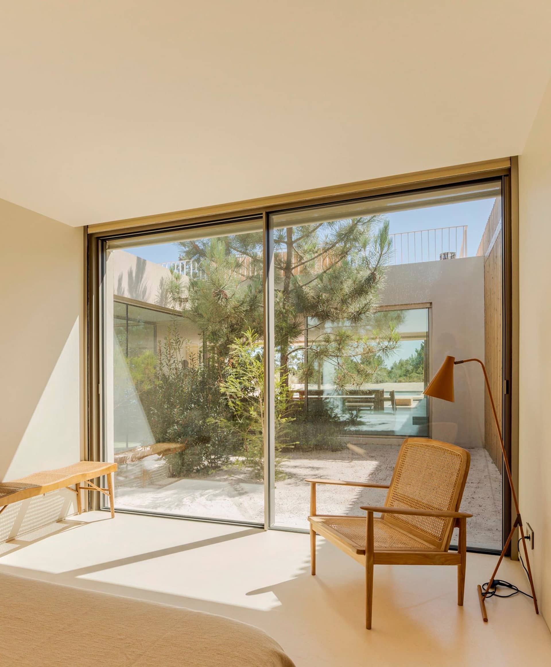 Modern bedroom with white flooring, wood furniture, and large glass sliding doors to an outdoor area with pines and a patio.