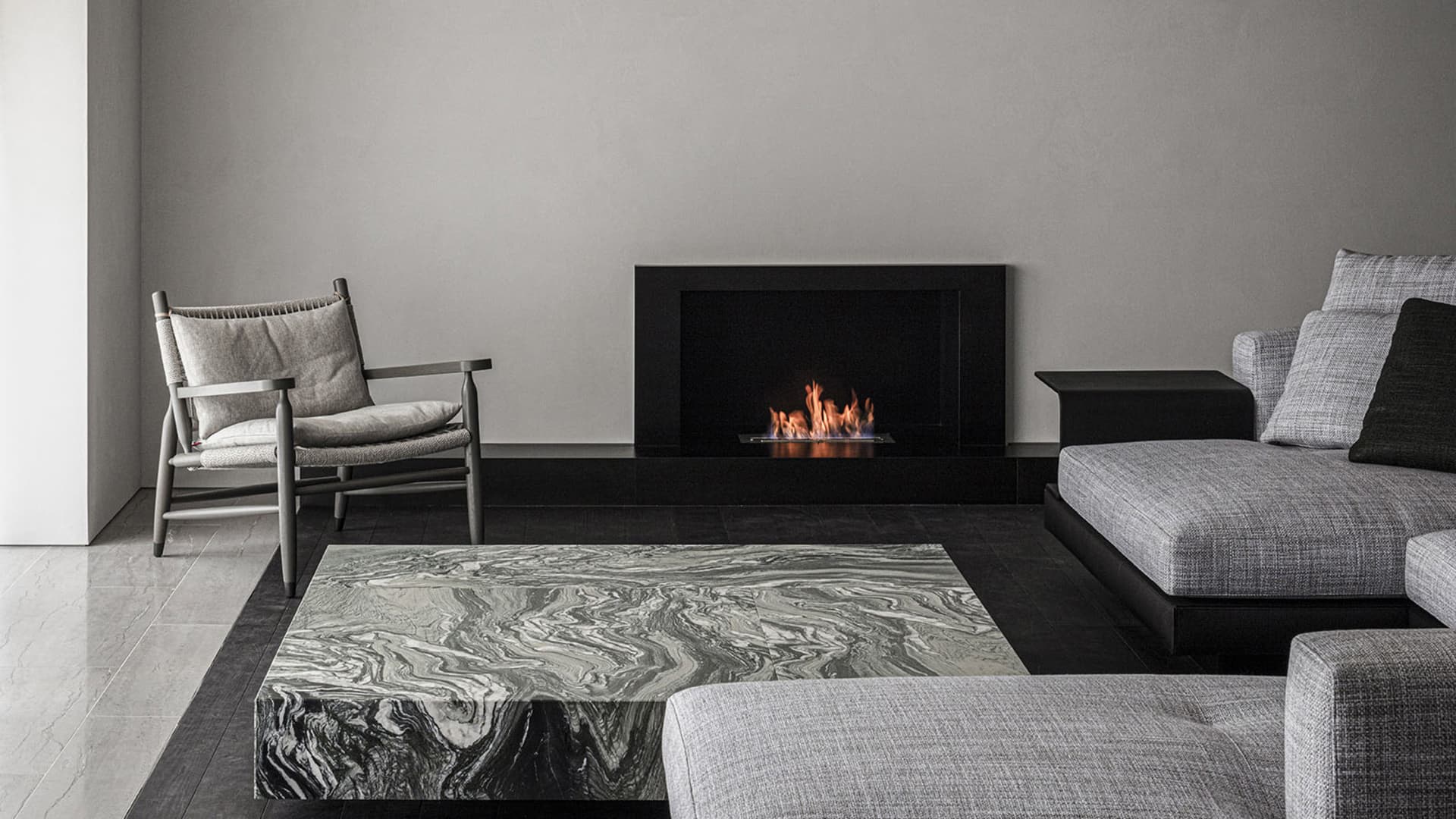 A modern minimalist living room featuring a fireplace, a marble coffee table, a grey sofa, and an armchair with cushions, all