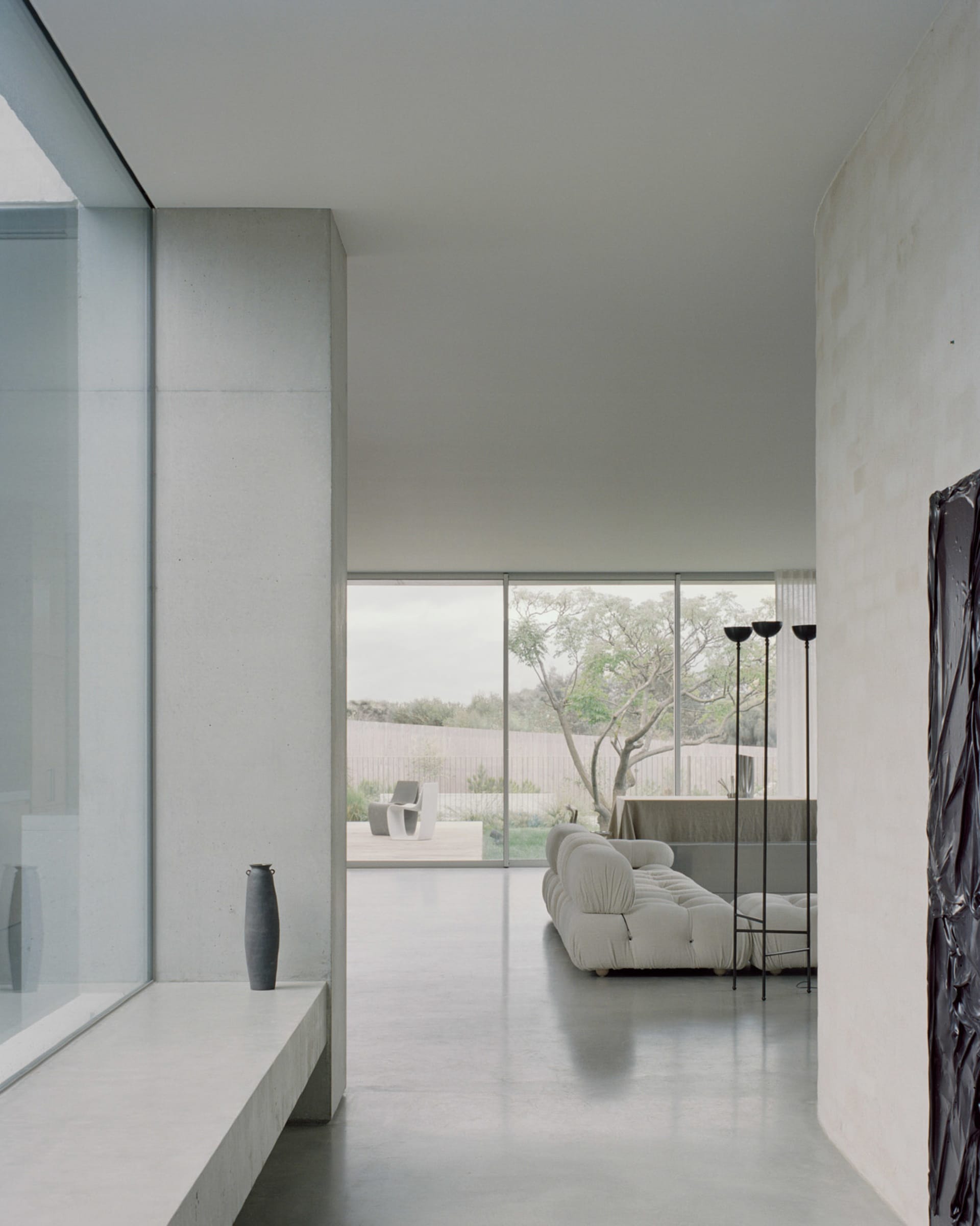 Modern, minimalist living room with concrete walls, a white sofa, and large windows offering a view of nature.
