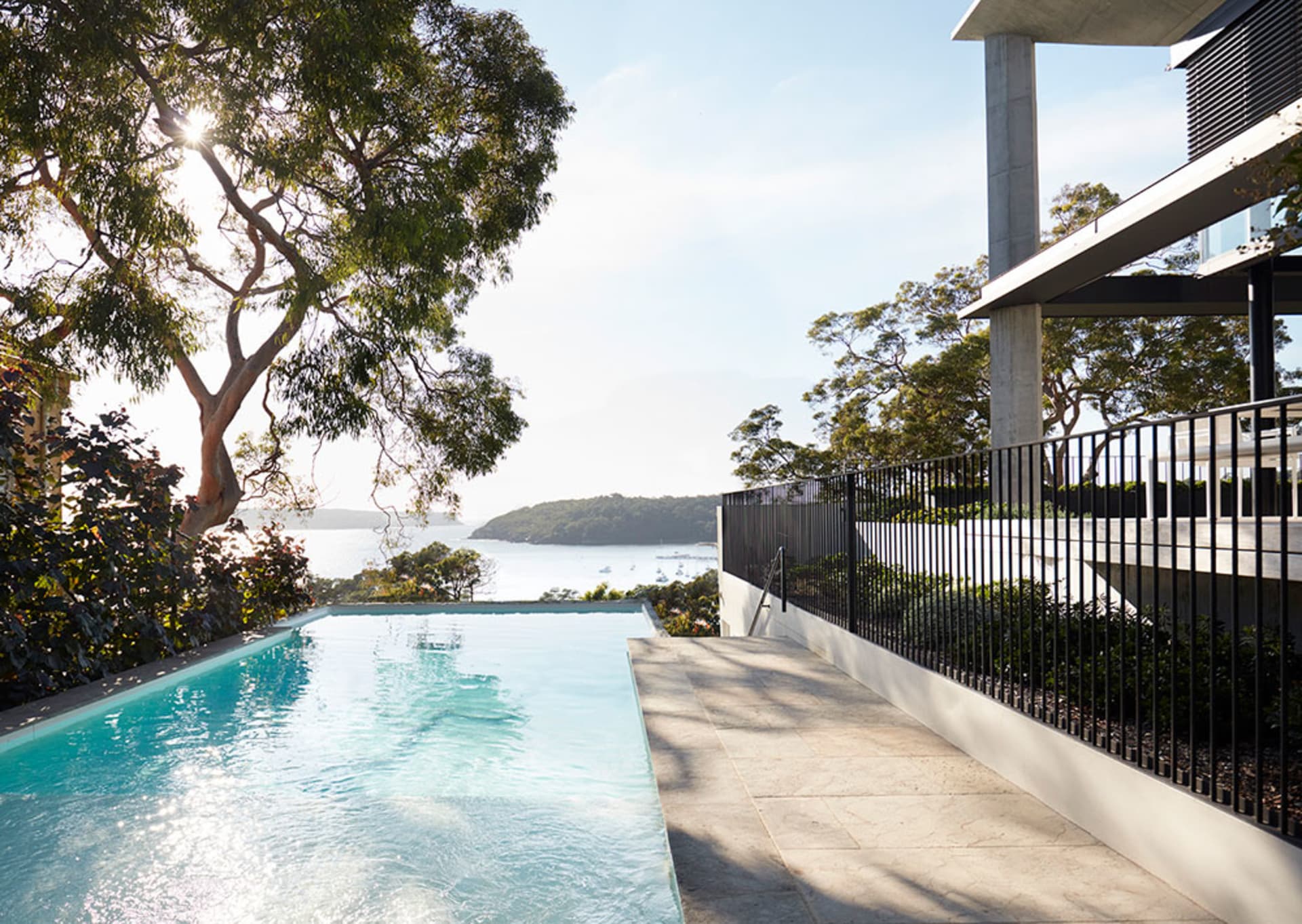 A modern outdoor residential area featuring a pool with a view of the sea, surrounded by greenery and a sleek, contemporary h