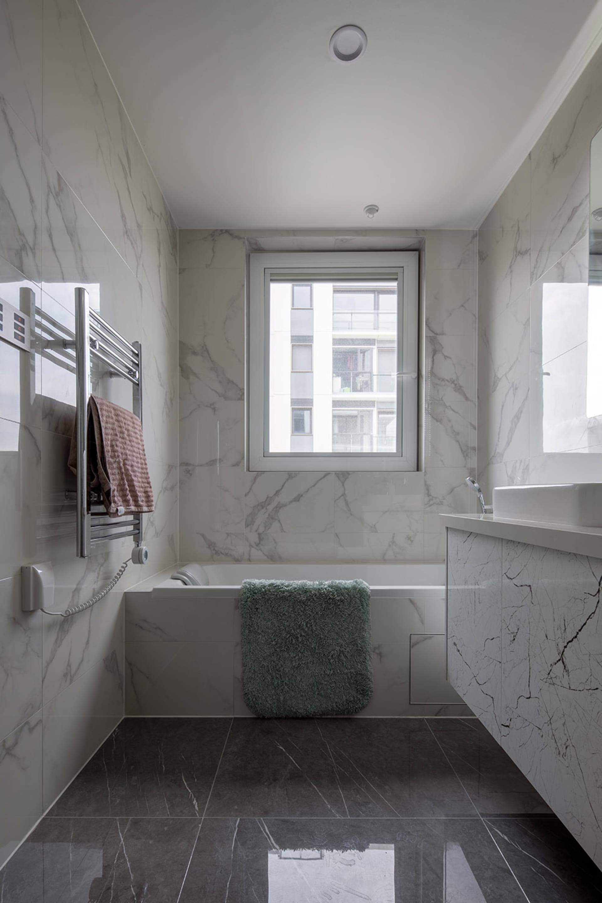 A minimalist bathroom with marble walls and floor, featuring a bathtub, sink, and towel rack.