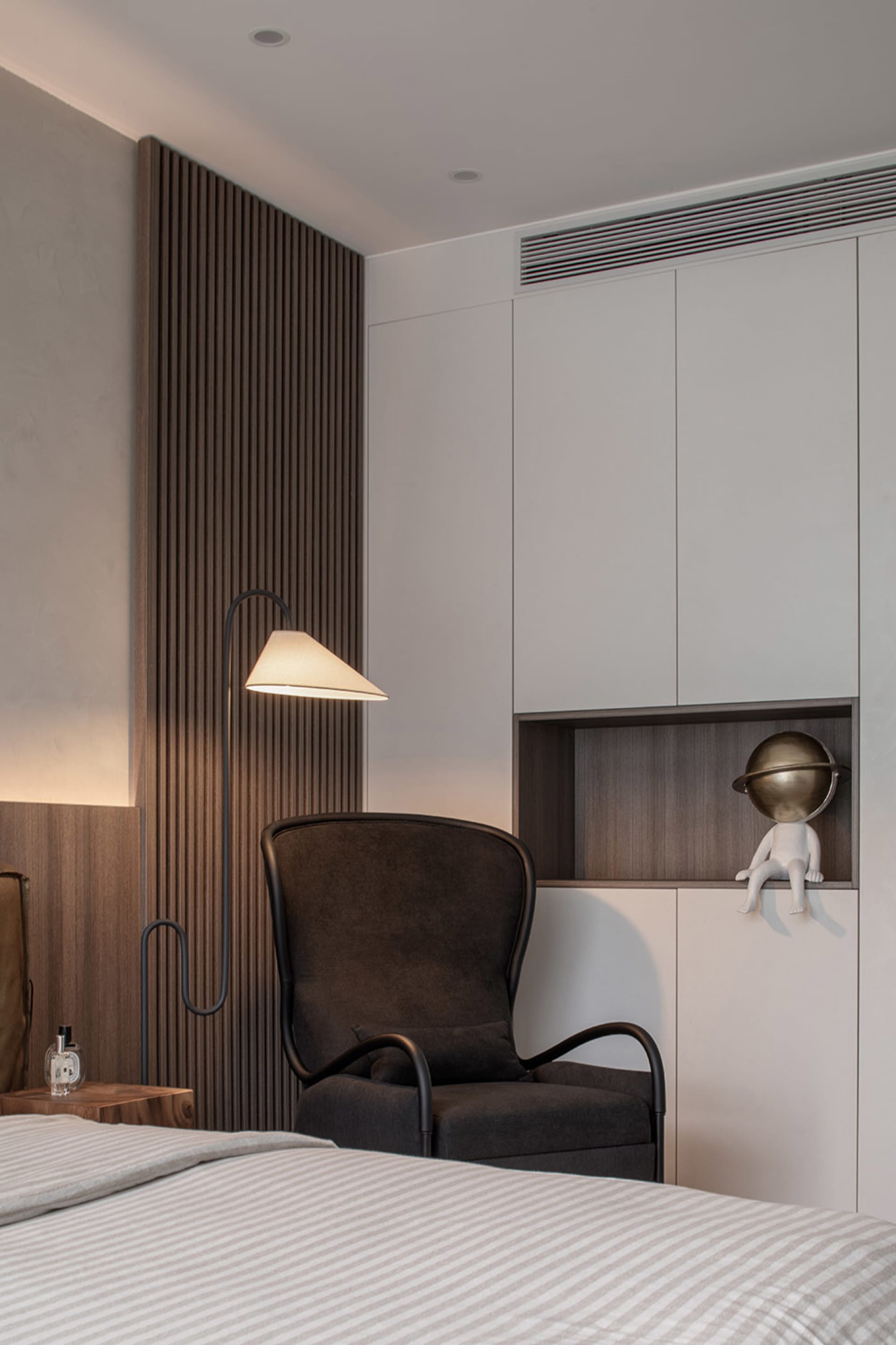 Modern bedroom with a dark wooden accent wall, a dark armchair, a floor lamp, and a white bed with striped sheets.