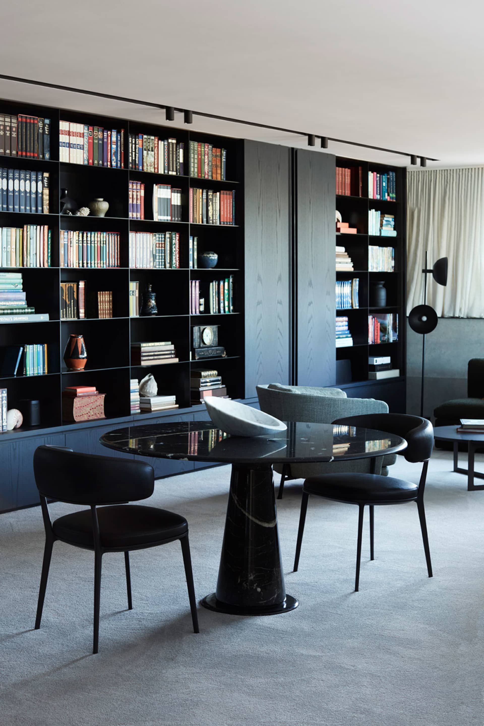 A modern library featuring marble bookshelves and a round black marble table with black leather chairs.