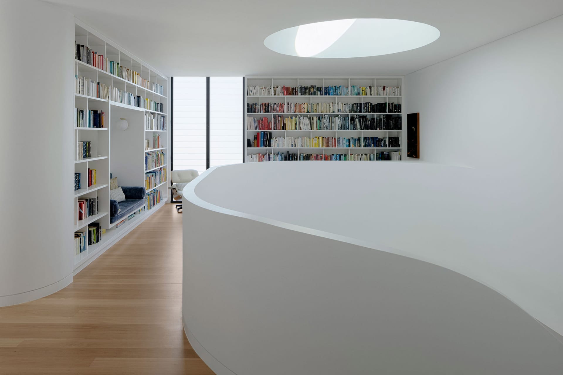 A modern library interior with white concrete walls, shelves filled with books, and an oval skylight.