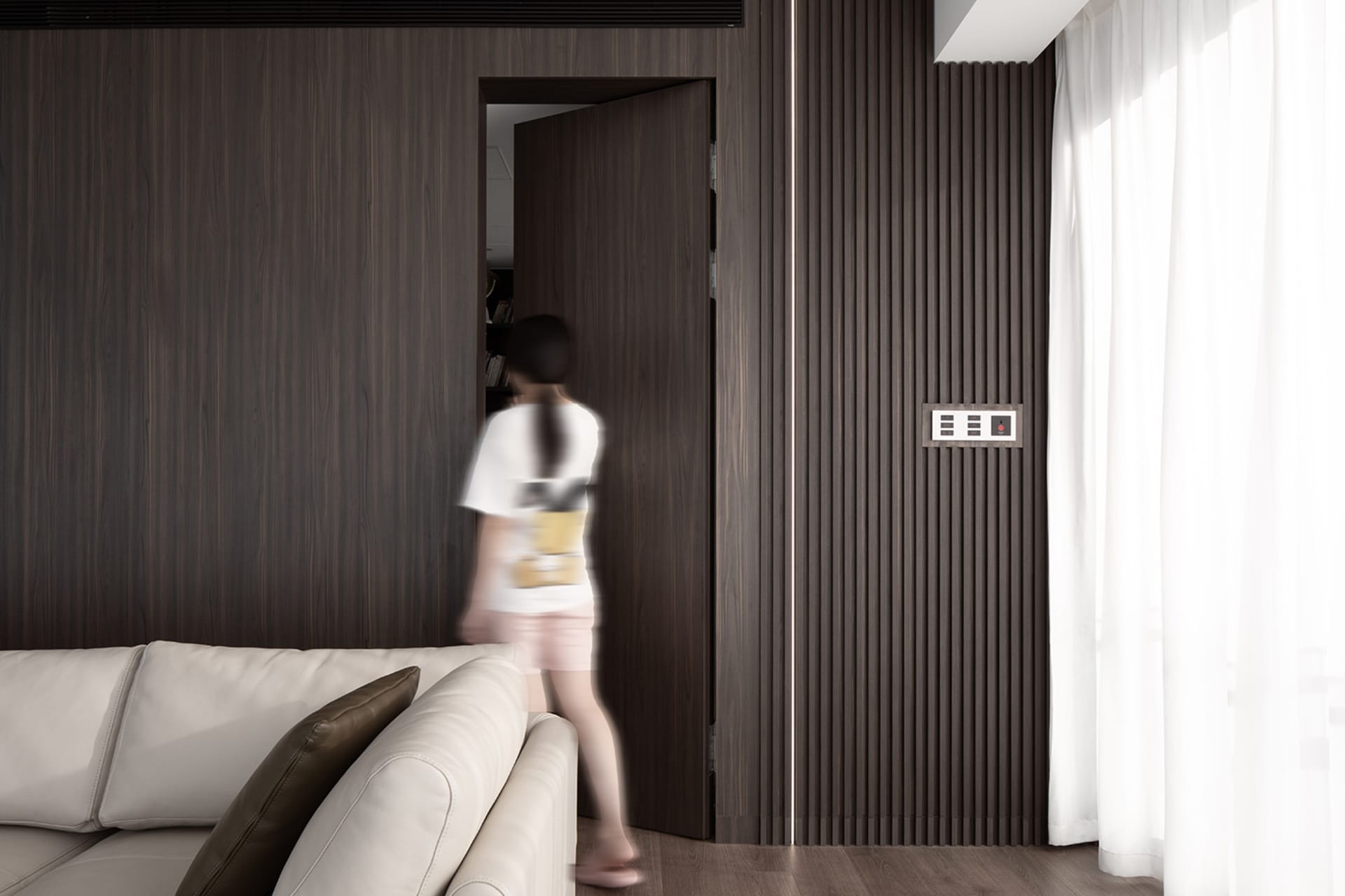 A modern interior design living room with wooden walls, a white sofa, and a woman walking through the doorway.