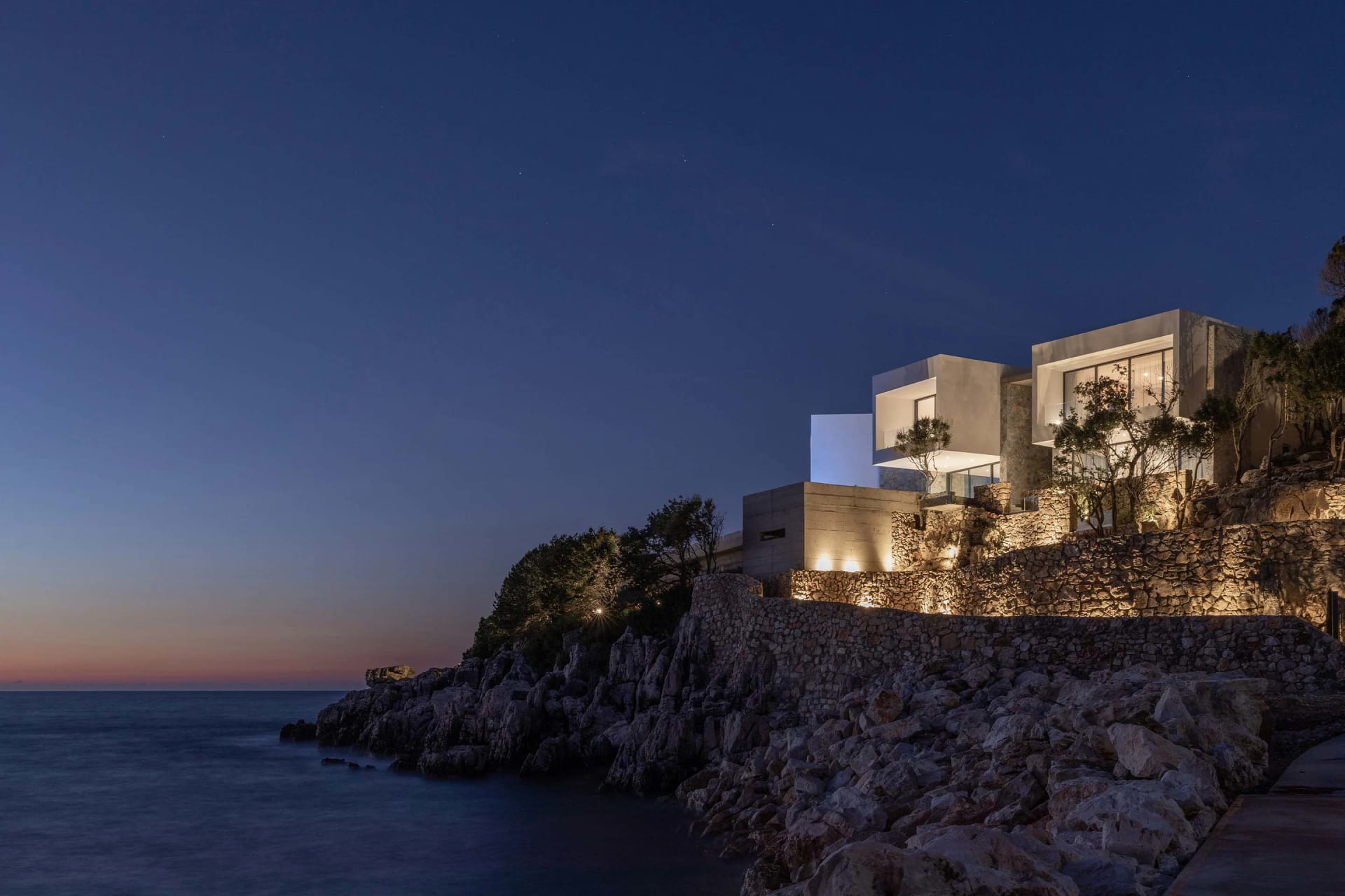 A modern house with glass walls and concrete structures by the ocean at dusk, illuminated by ambient lighting.