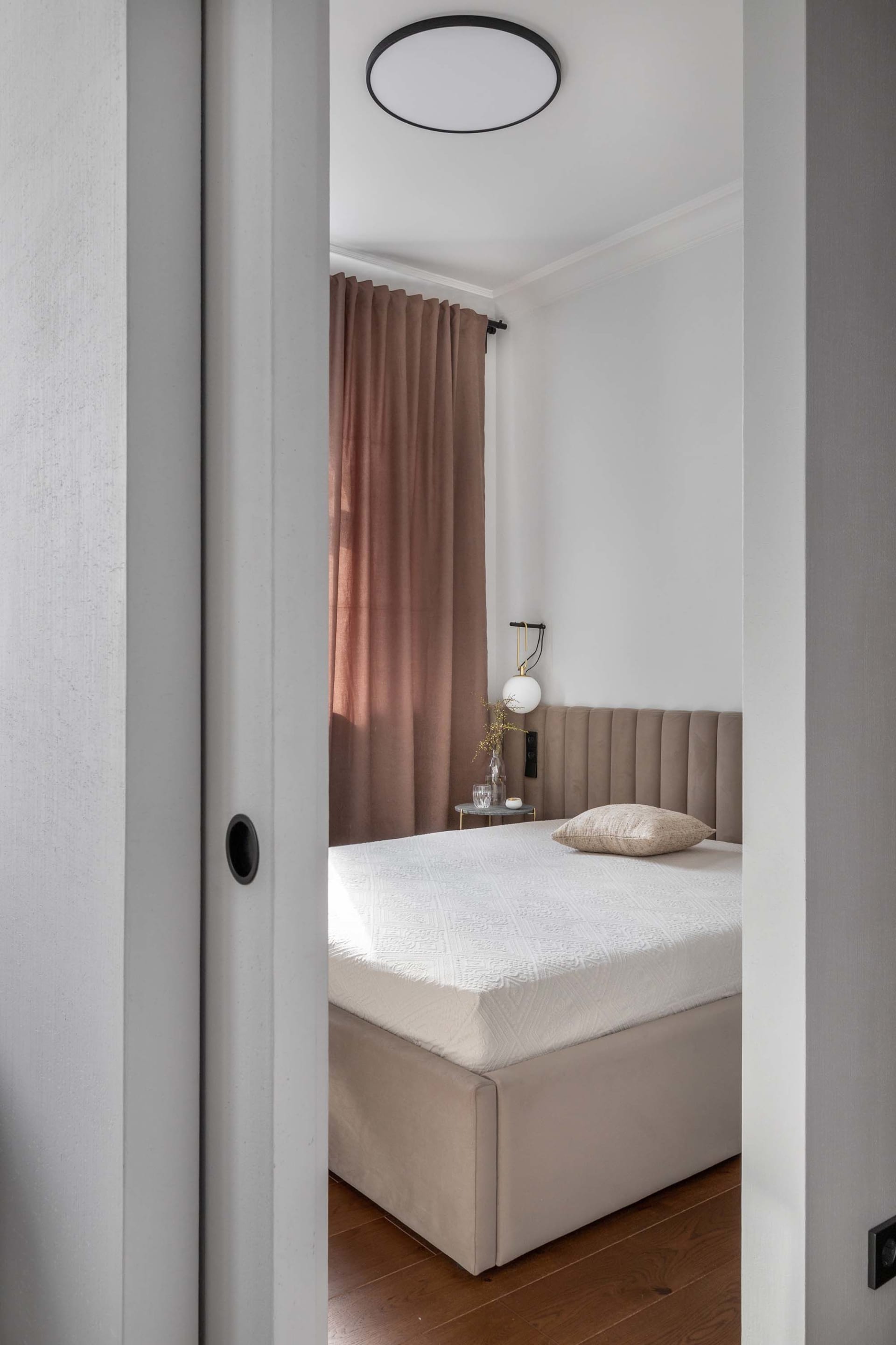 A minimalist bedroom with a beige upholstered bed, brown velvet curtains, and wooden floor.