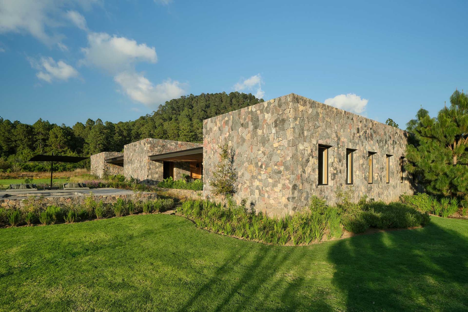 A modern stone house with a peaceful, green landscape and a forested hill in the background.