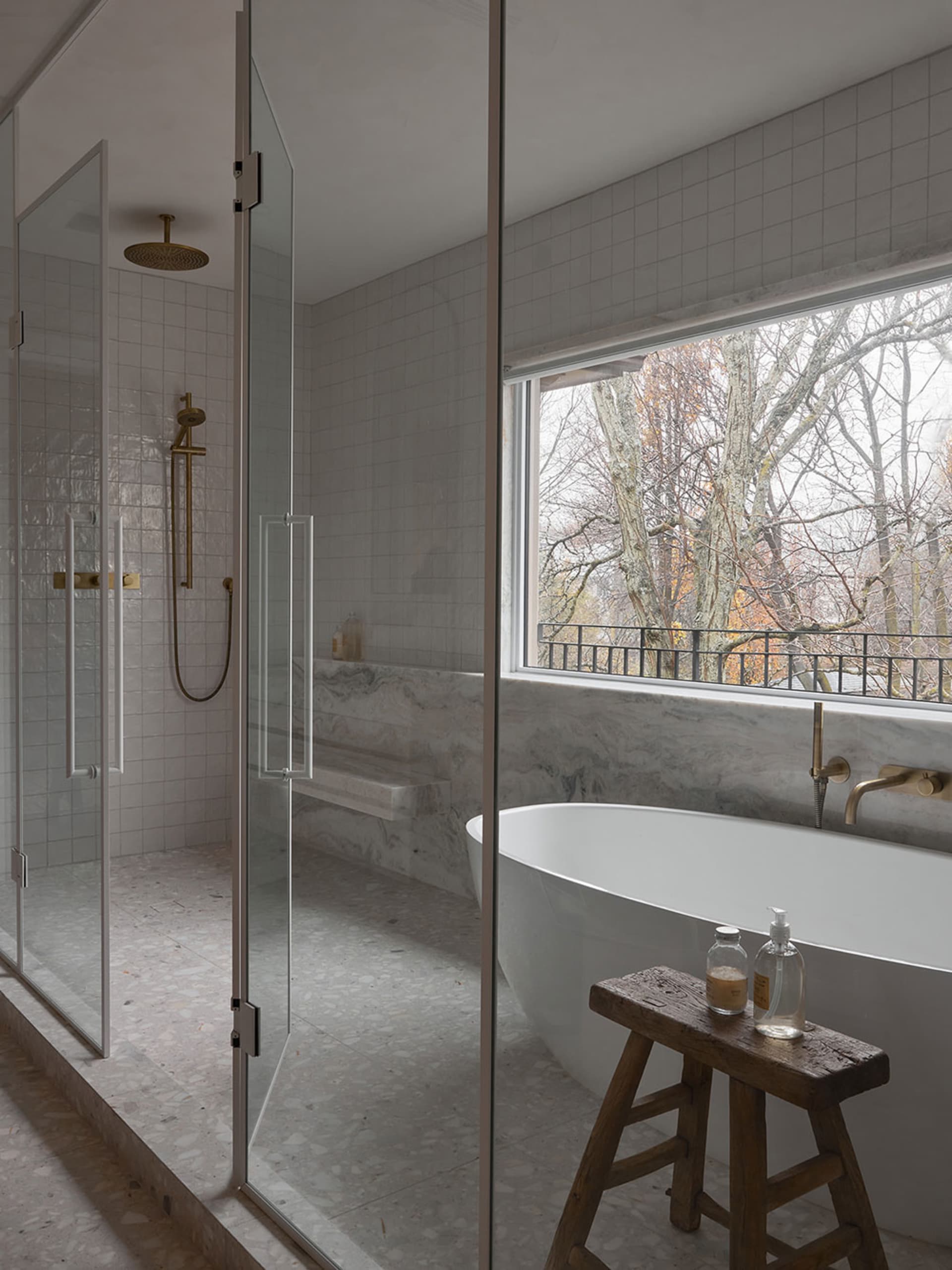 Modern minimalist bathroom featuring a white marble tub, glass shower enclosure, and a window overlooking trees.
