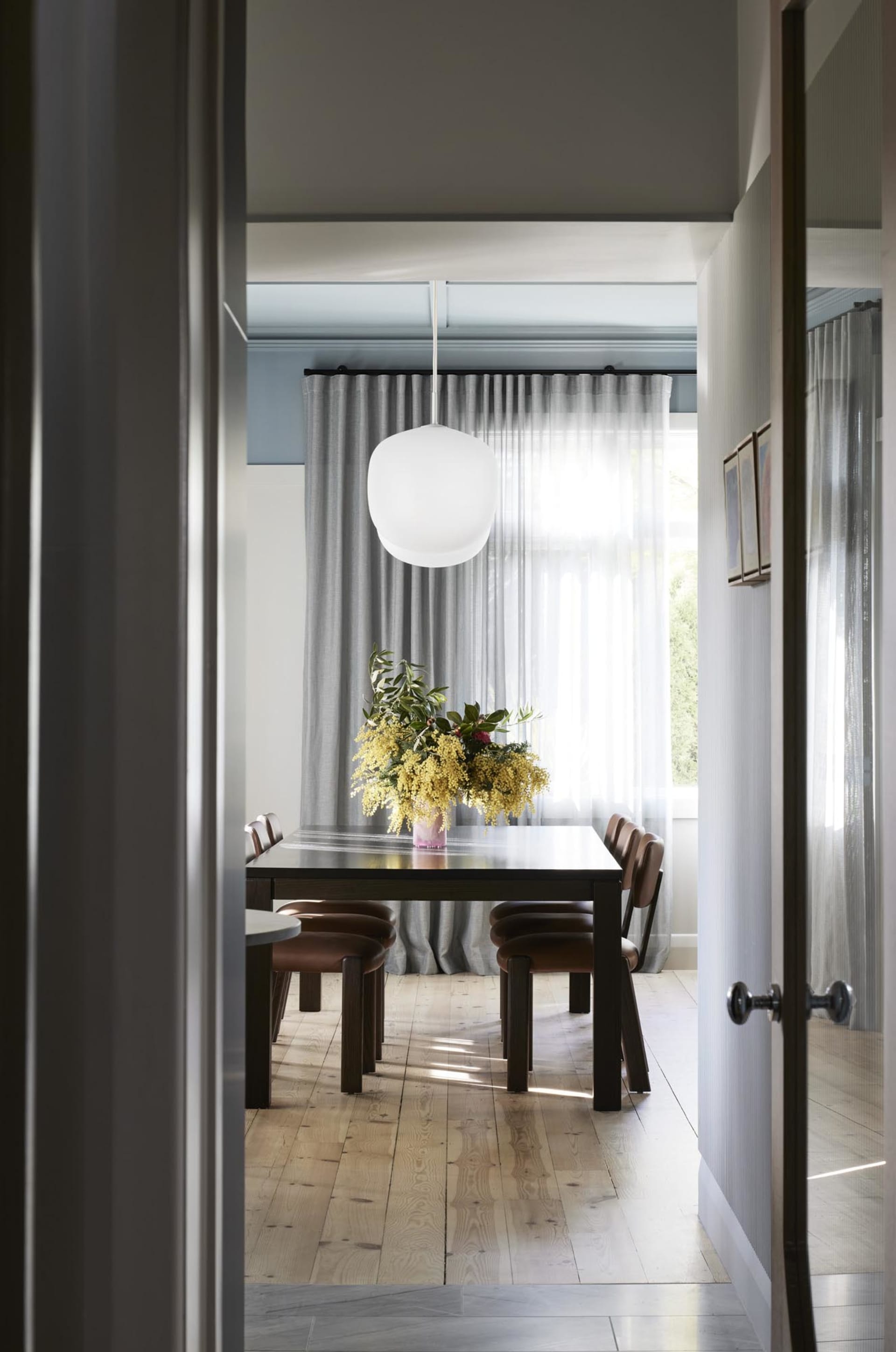 A modern dining room with a wooden table and leather chairs, featuring a white pendant light and sheer curtains.