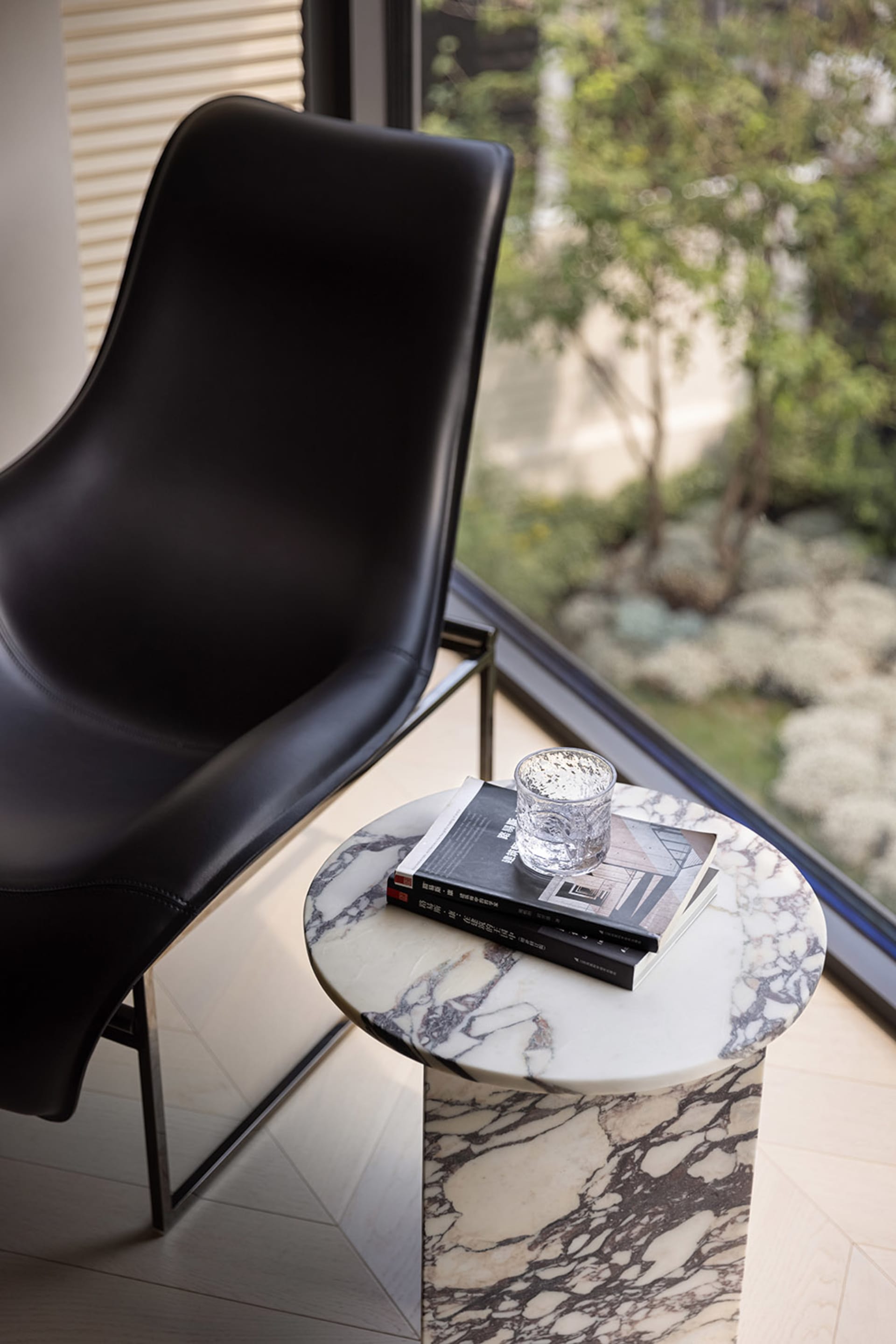 A minimalist modern penthouse with a sleek black chair, marble side table, and a glass of water, featuring a view of greenery