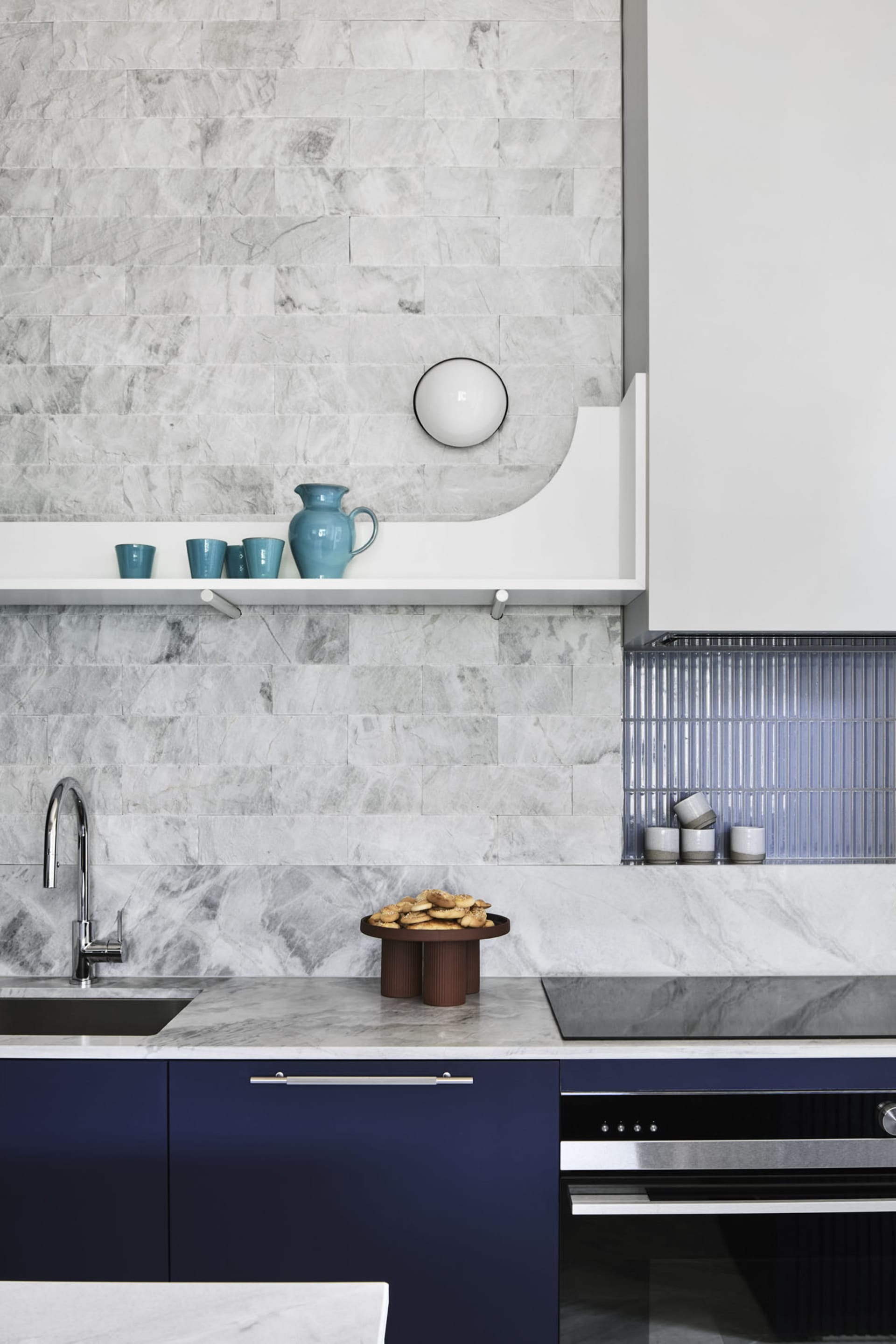 A modern kitchen with marble countertops and a blue cabinet beneath, featuring a marble backsplash, blue cups on a shelf, and