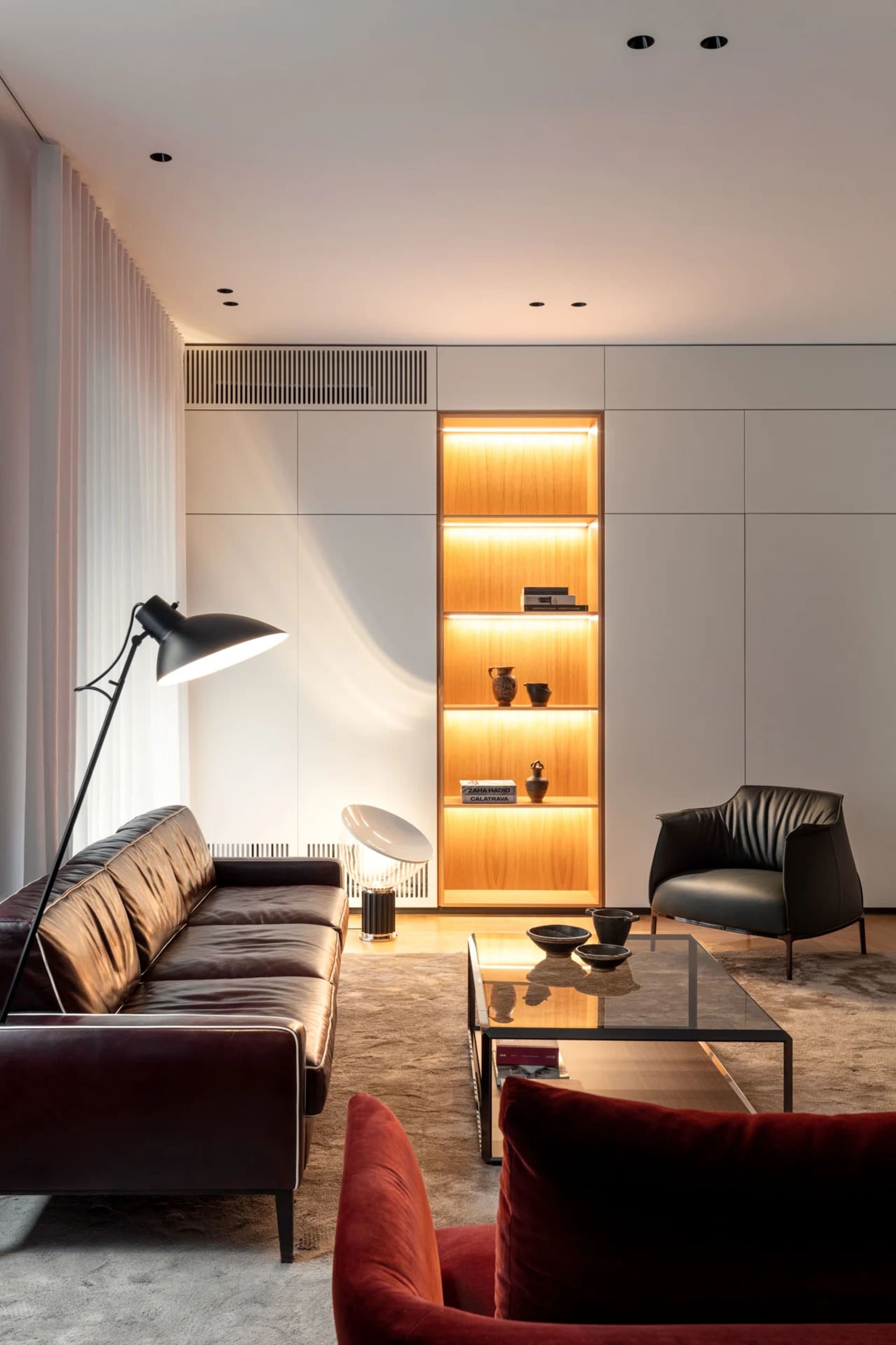 A modern living room featuring a large leather sofa, a glass coffee table, and a tall wooden shelf with illuminated lighting.