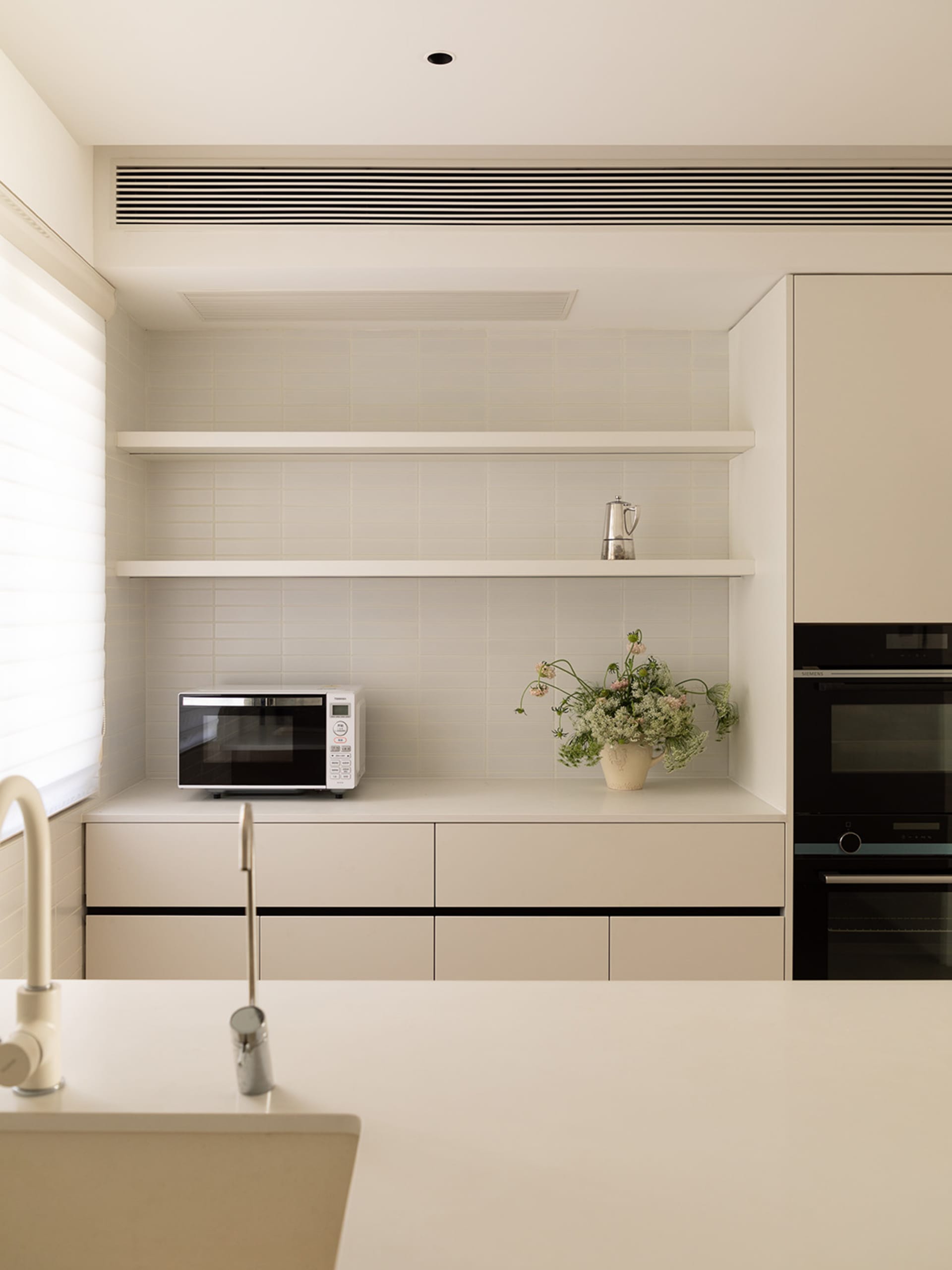 A modern, minimalist residential kitchen with white cabinetry, shelves, and appliances, featuring a vase of flowers and a mic