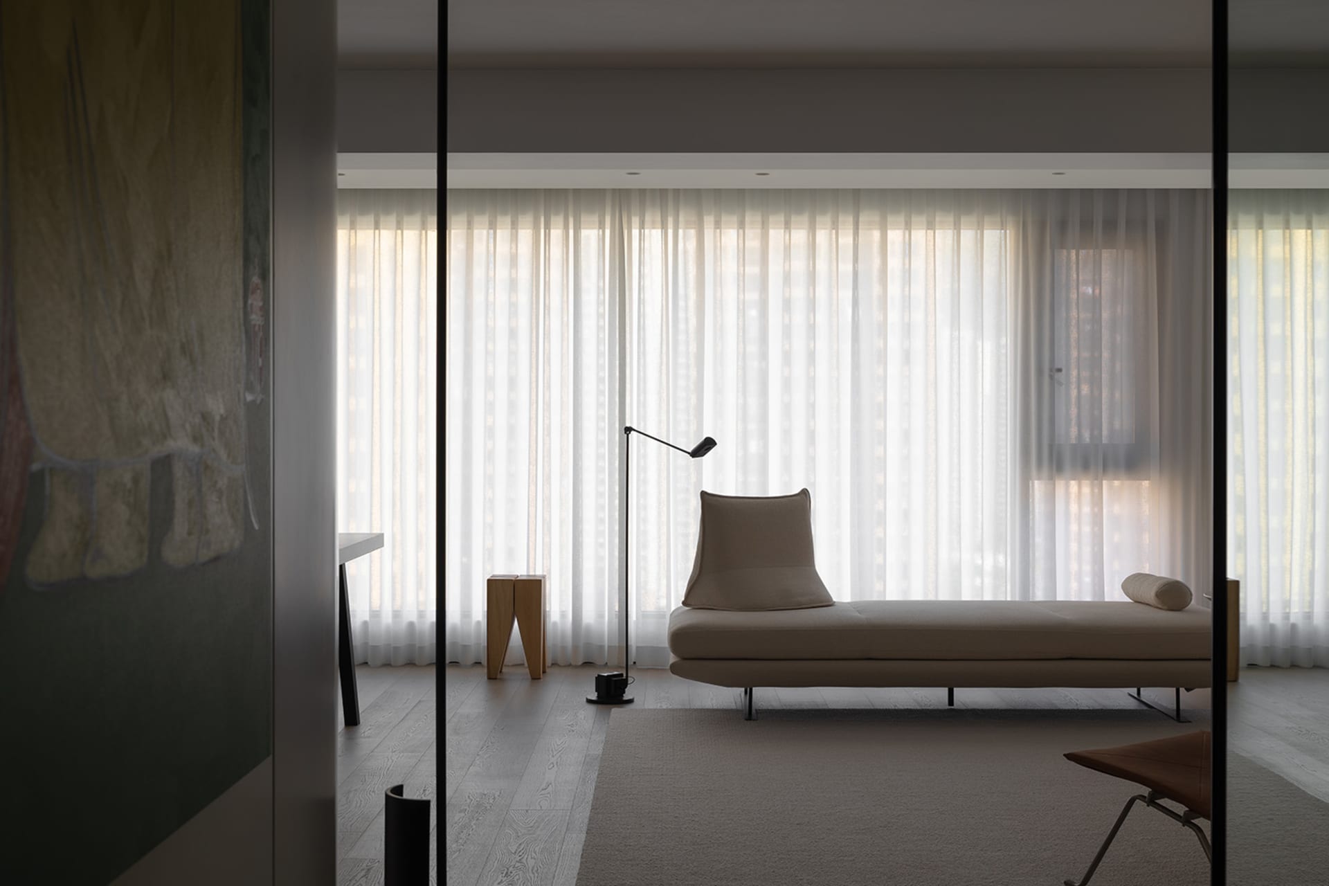 A minimalist residential lounge with a beige chaise lounge, a floor lamp, and sheer curtains allowing light to filter in.