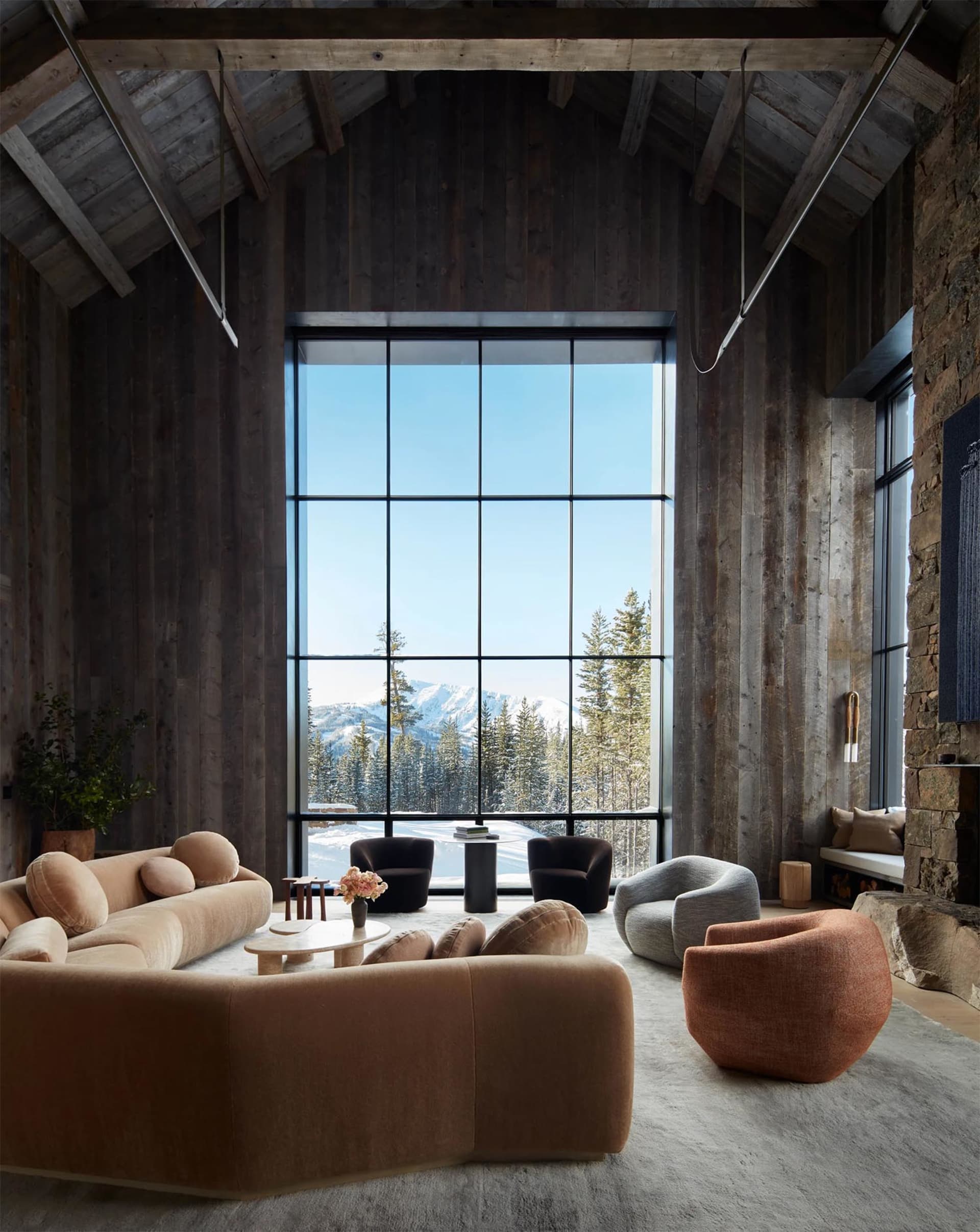 Modern living room with large windows overlooking snowy mountains, featuring a comfortable sofa, armchairs, and a stone firep