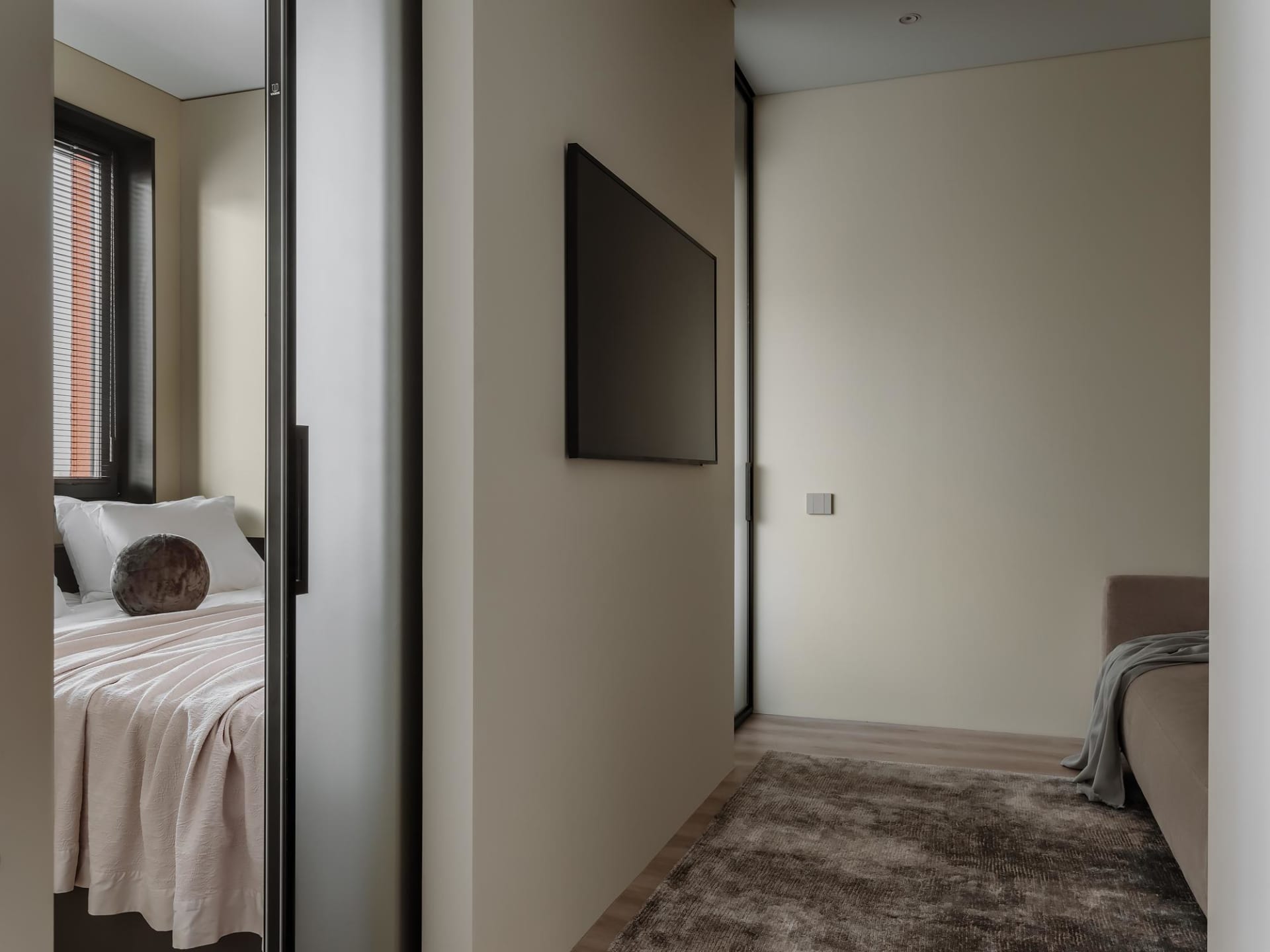 A modern bedroom with neutral-colored walls, a large flat-screen TV mounted on the wall, and a minimalistic design.