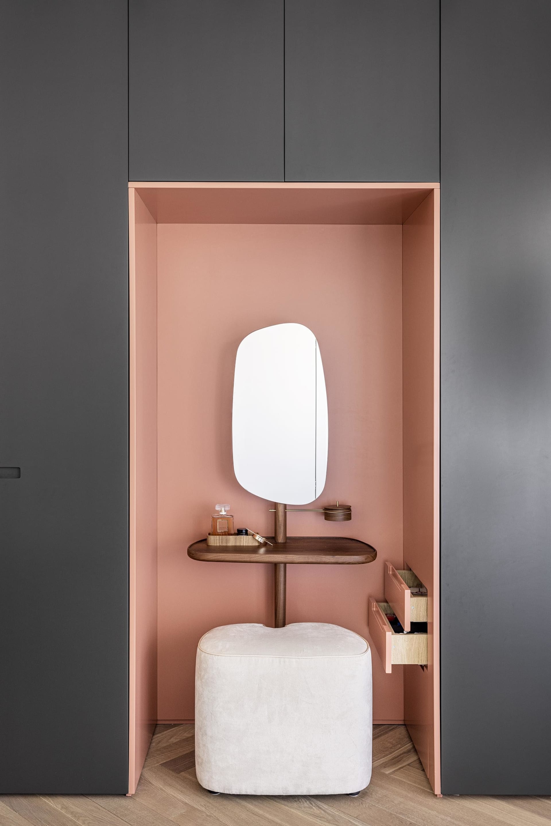 Minimalist vanity area with a white stool, pink walls, and a white shelf holding a perfume bottle and a brush.
