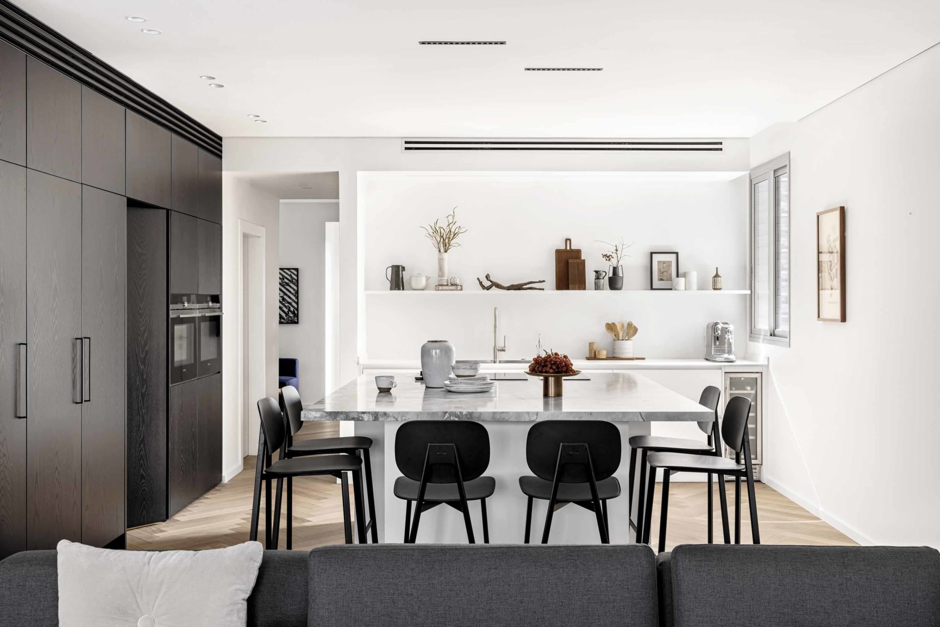 A modern kitchen featuring a black marble island with black chairs and wooden flooring, surrounded by white walls and black c