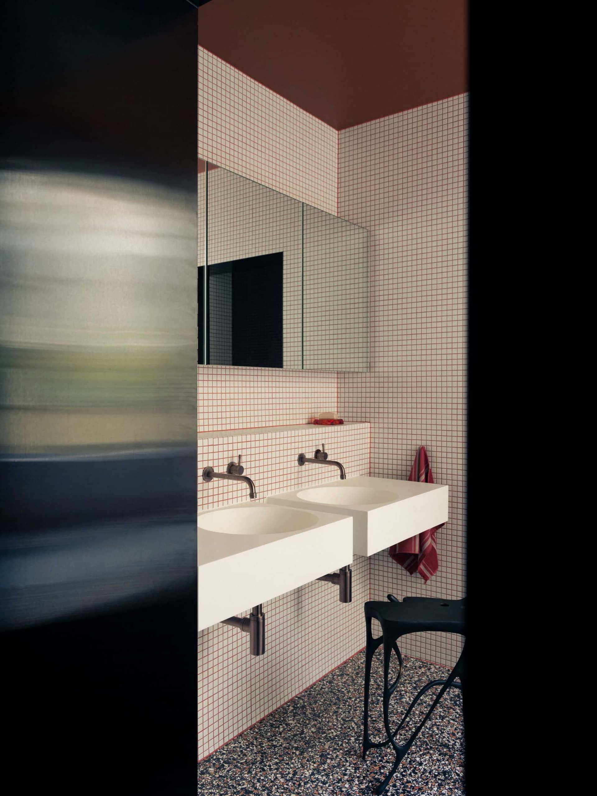 Modern bathroom with white and red tiled walls, white countertops, and black metal stool with a terrazzo floor.