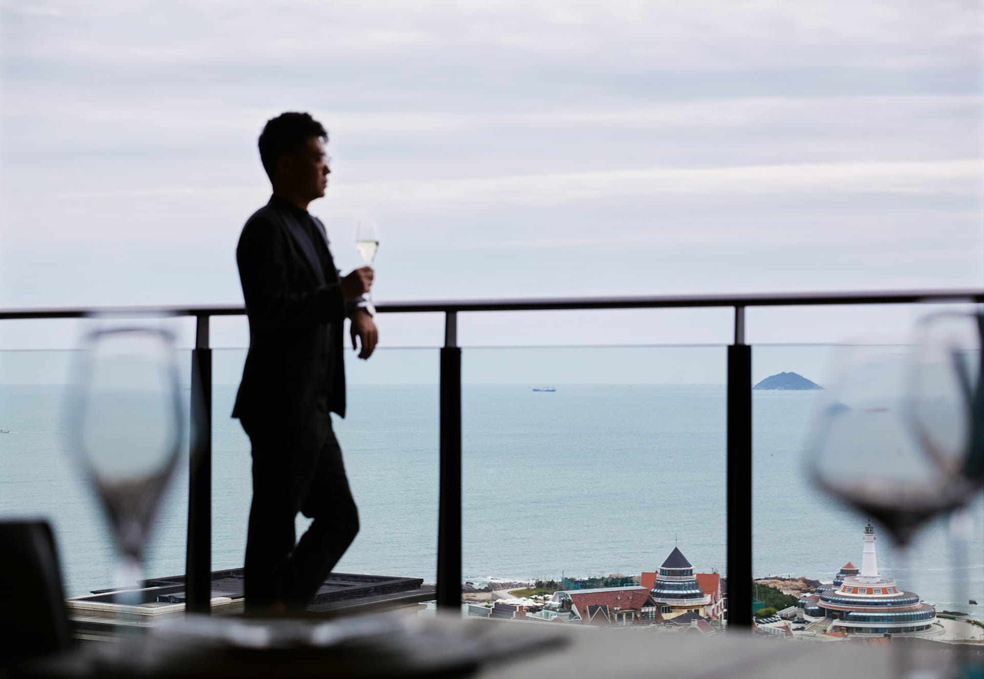 男子站在陽台，手持紅酒杯，眺望大海。