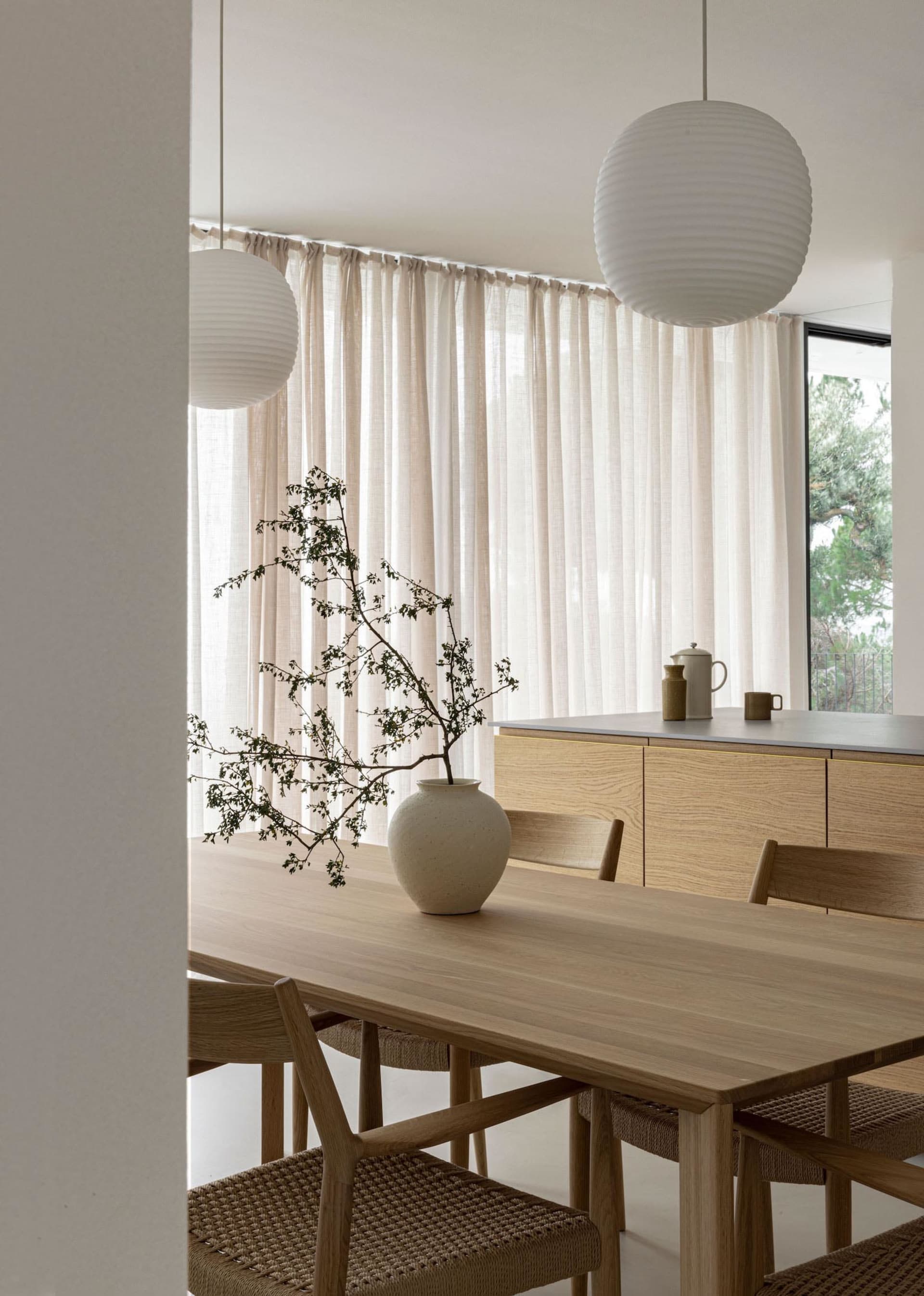 A minimalist dining room featuring a wooden table with a vase of branches, white pendant lights, and sheer curtains, with a n