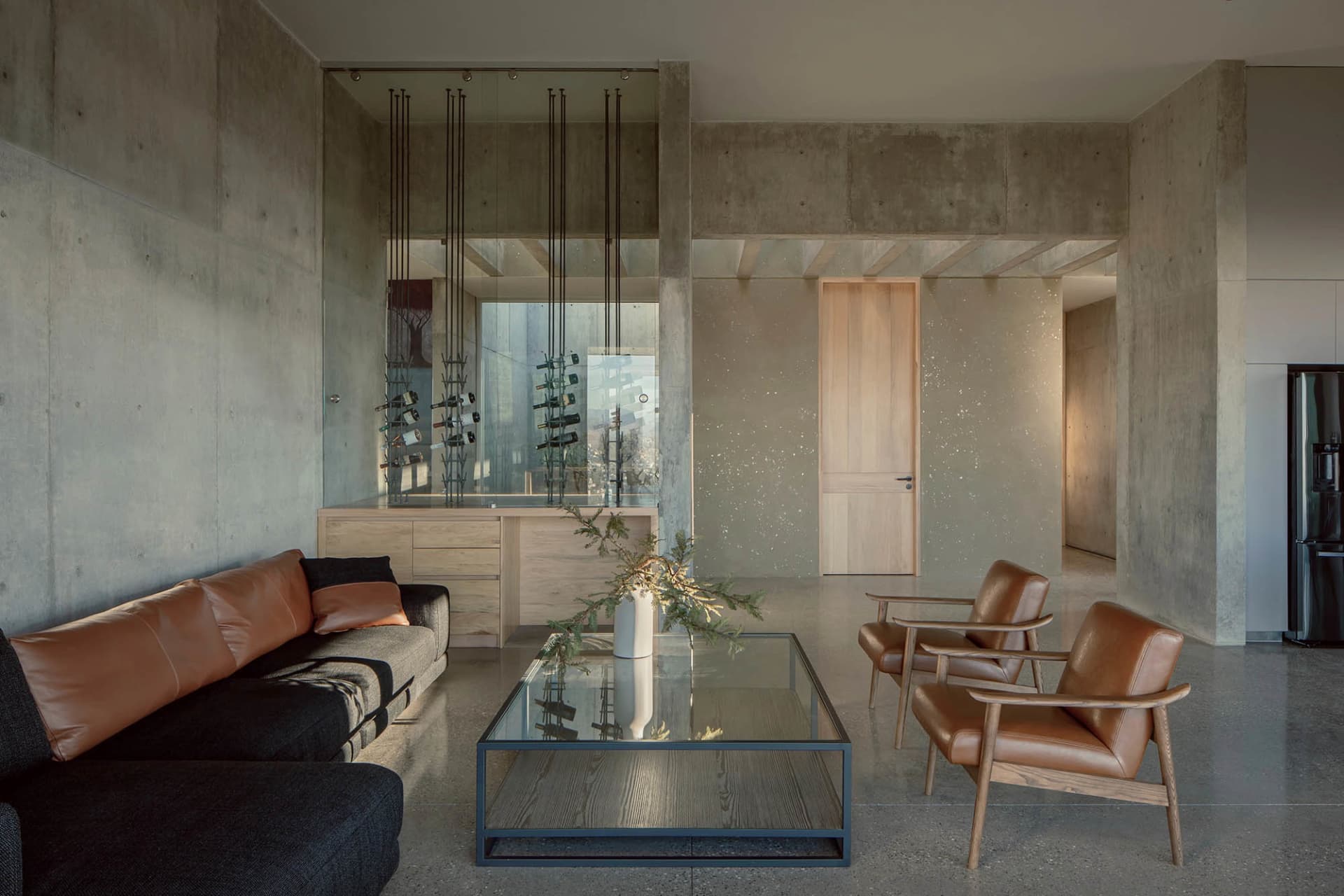 Modern industrial living room with concrete walls, a glass coffee table, leather armchairs, and a wine rack.