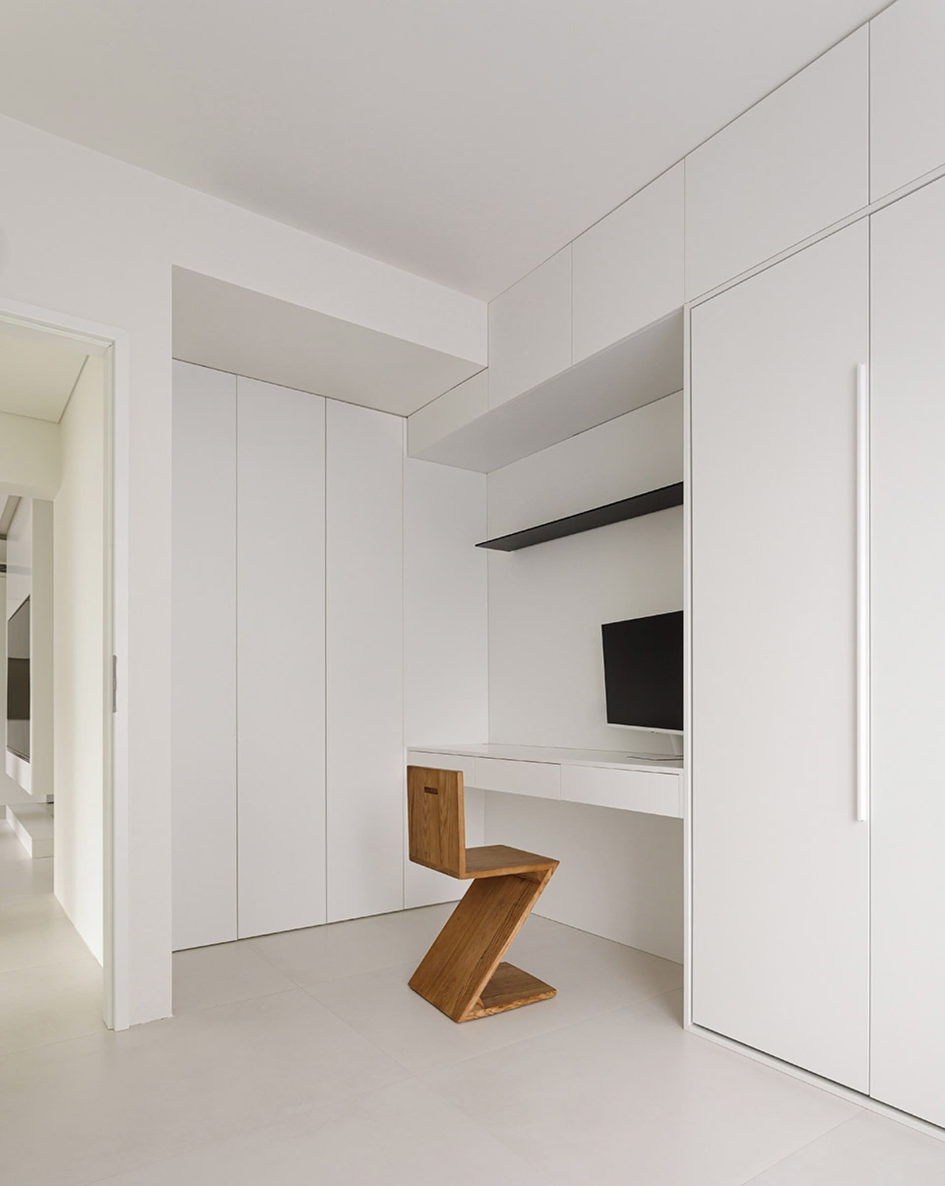 A minimalist workspace with a white floor and walls, featuring a wooden Z-shaped chair and a built-in desk.