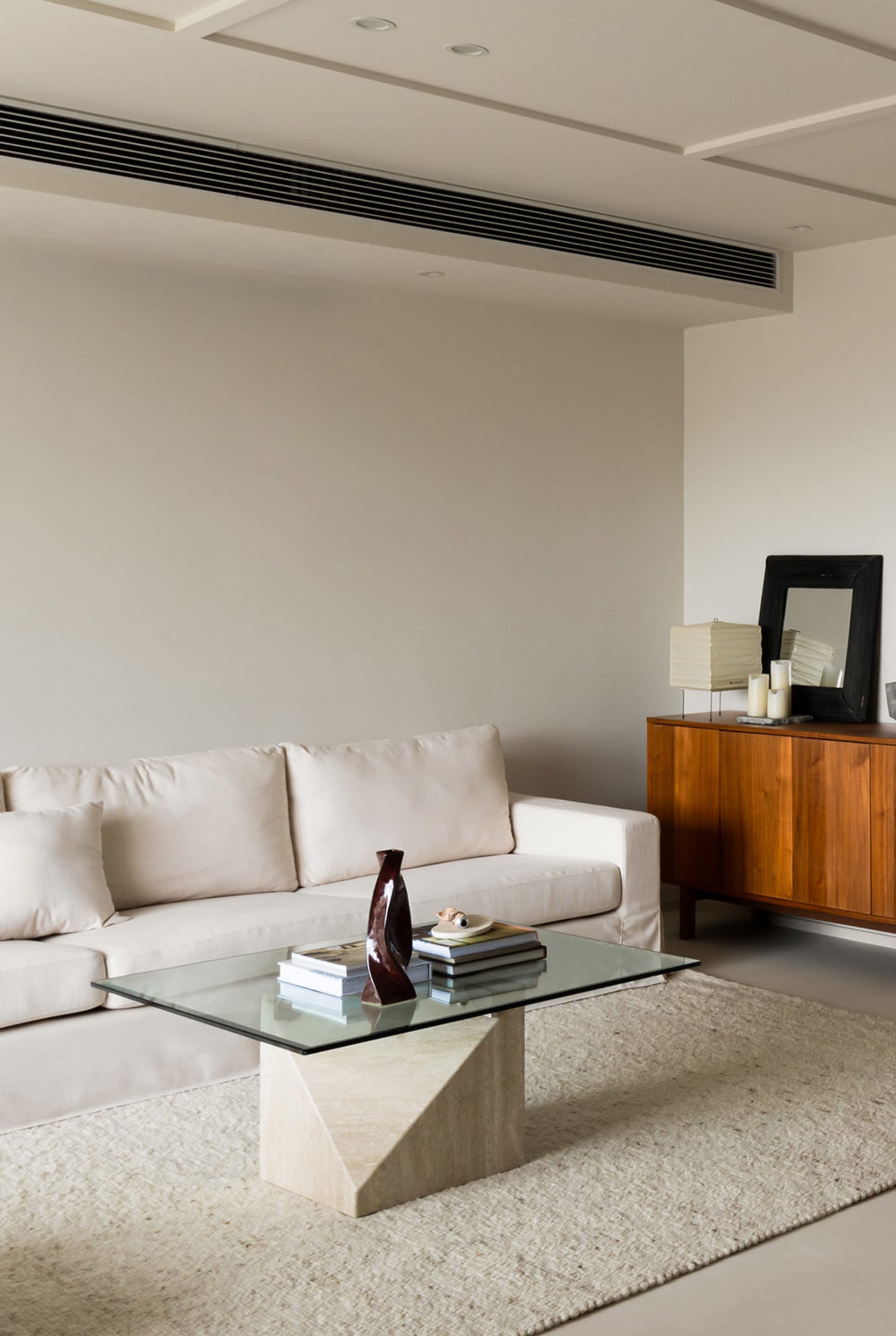 A modern living room with minimalist furniture and neutral-toned decor, featuring a glass-top coffee table, a wooden sideboar